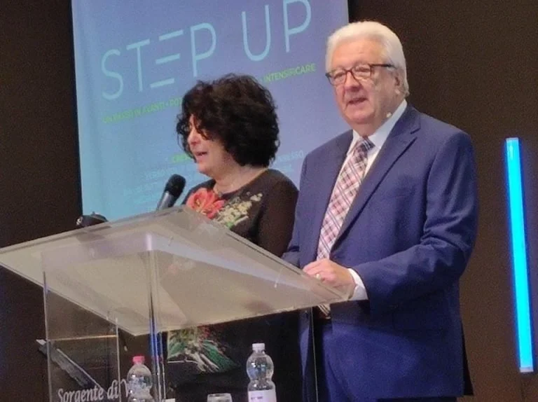 A man and woman standing at a podium speaking at an event, with a large screen behind them displaying the words 'STEP UP' and other text.