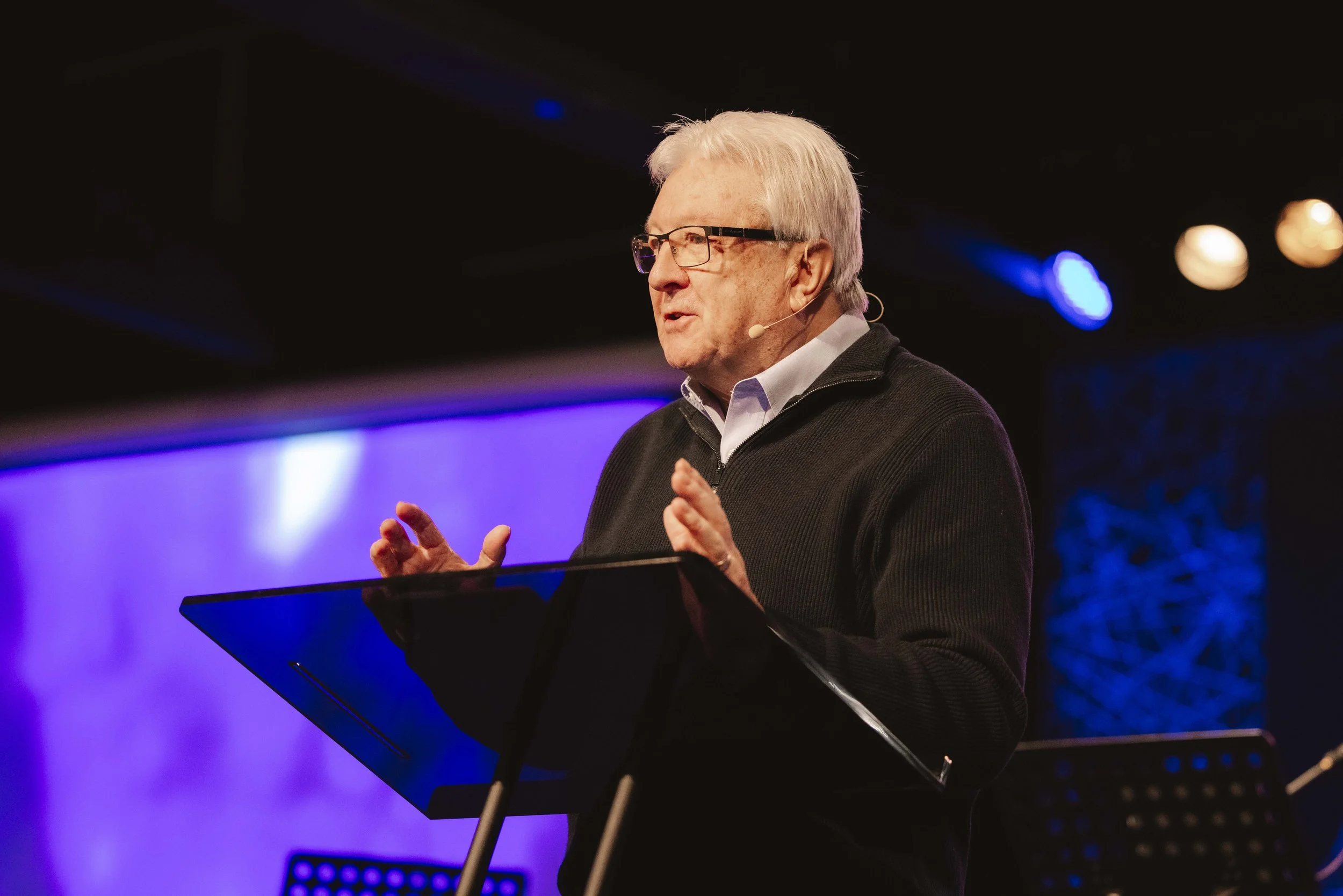 An elderly man with glasses speaking at a podium with a microphone attached, on a stage illuminated with purple and blue lighting.