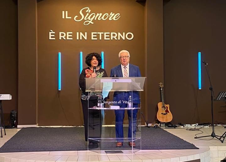 A woman and a man are standing behind a transparent lectern on a stage, delivering a speech or sermon. The background has a large sign with Italian text that reads 'IL Signore È RE IN ETERNO.' There is an acoustic guitar on the right side and some mu