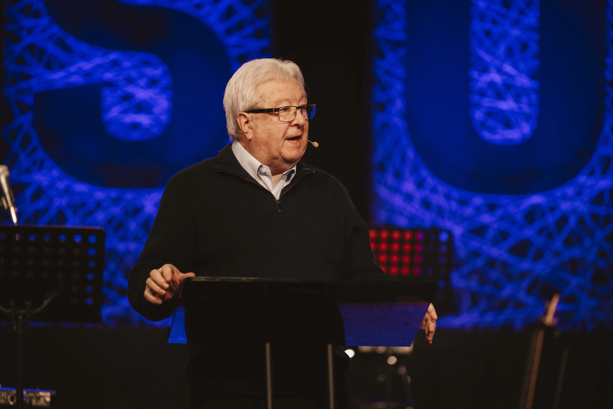 An elderly man with glasses and white hair giving a speech or lecture on stage, with a microphone headset, in front of an abstract background of blue and purple lights.