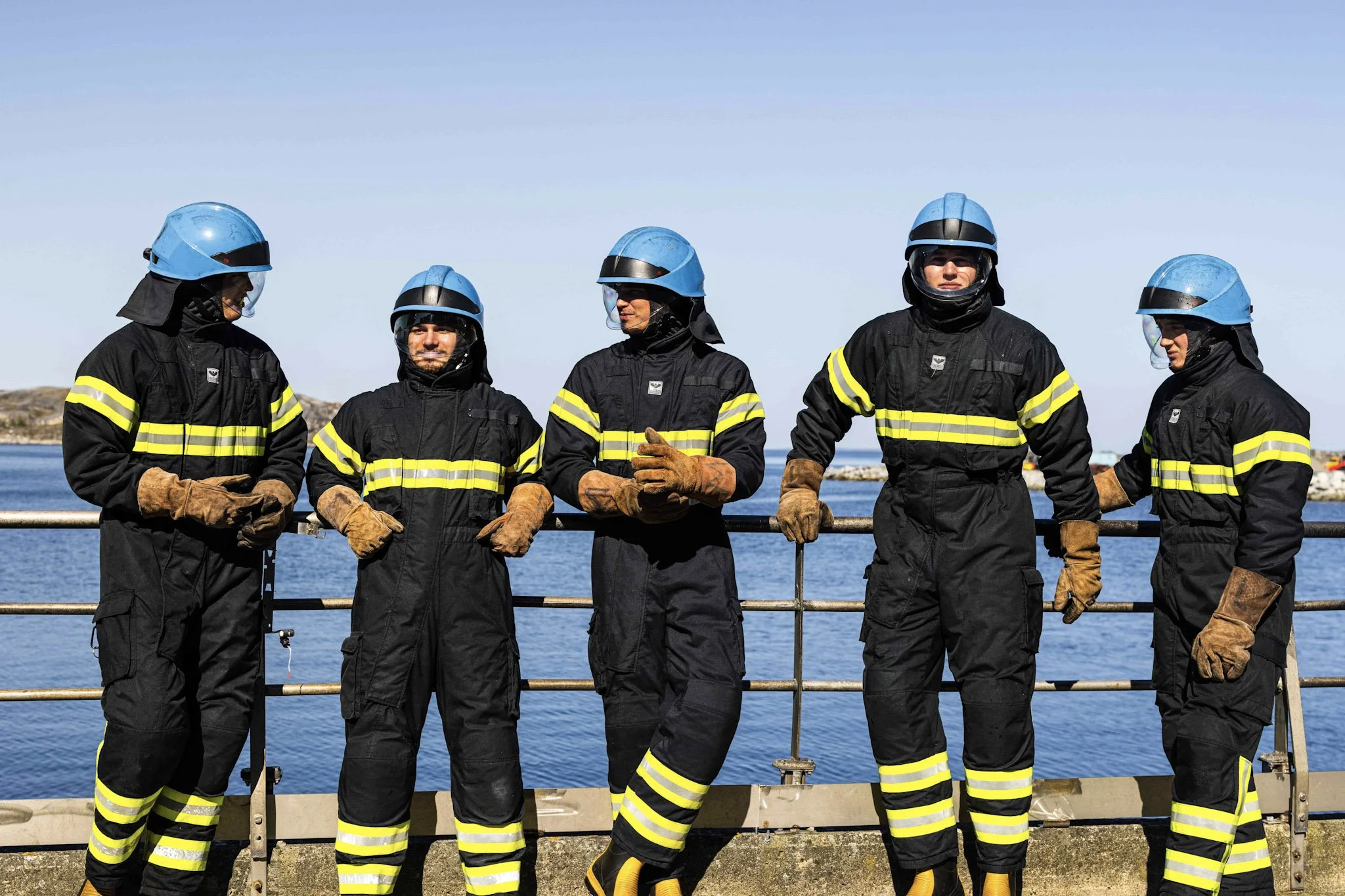 Fem brannmenn i svart og gult verneutstyr og hjelm står på en kai med vann i bakgrunnen.