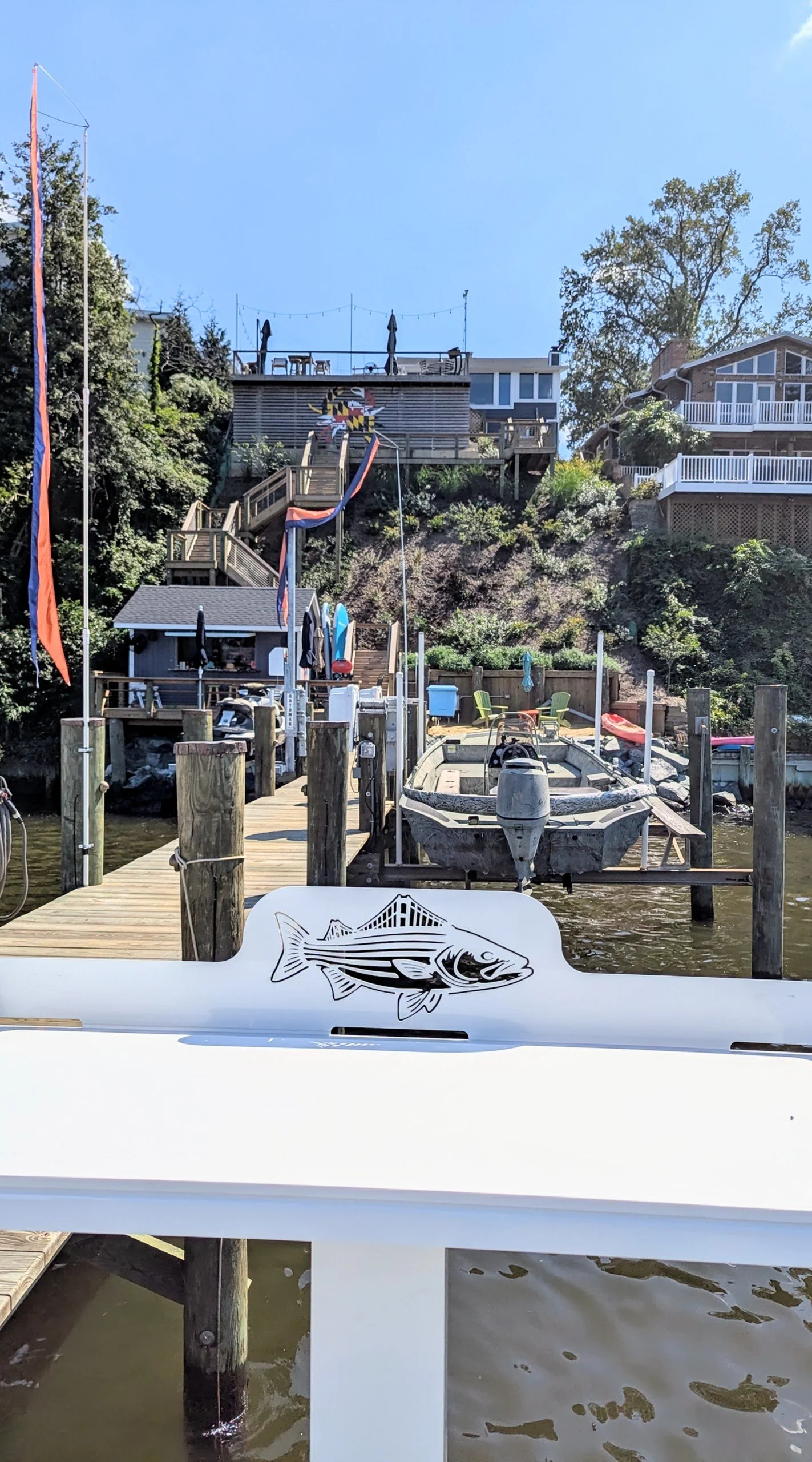 View of a dock on a waterway with boats, a wooden house with a large wooden crab sculpture painted with the Maryland flag, and houses on a hillside in the background.