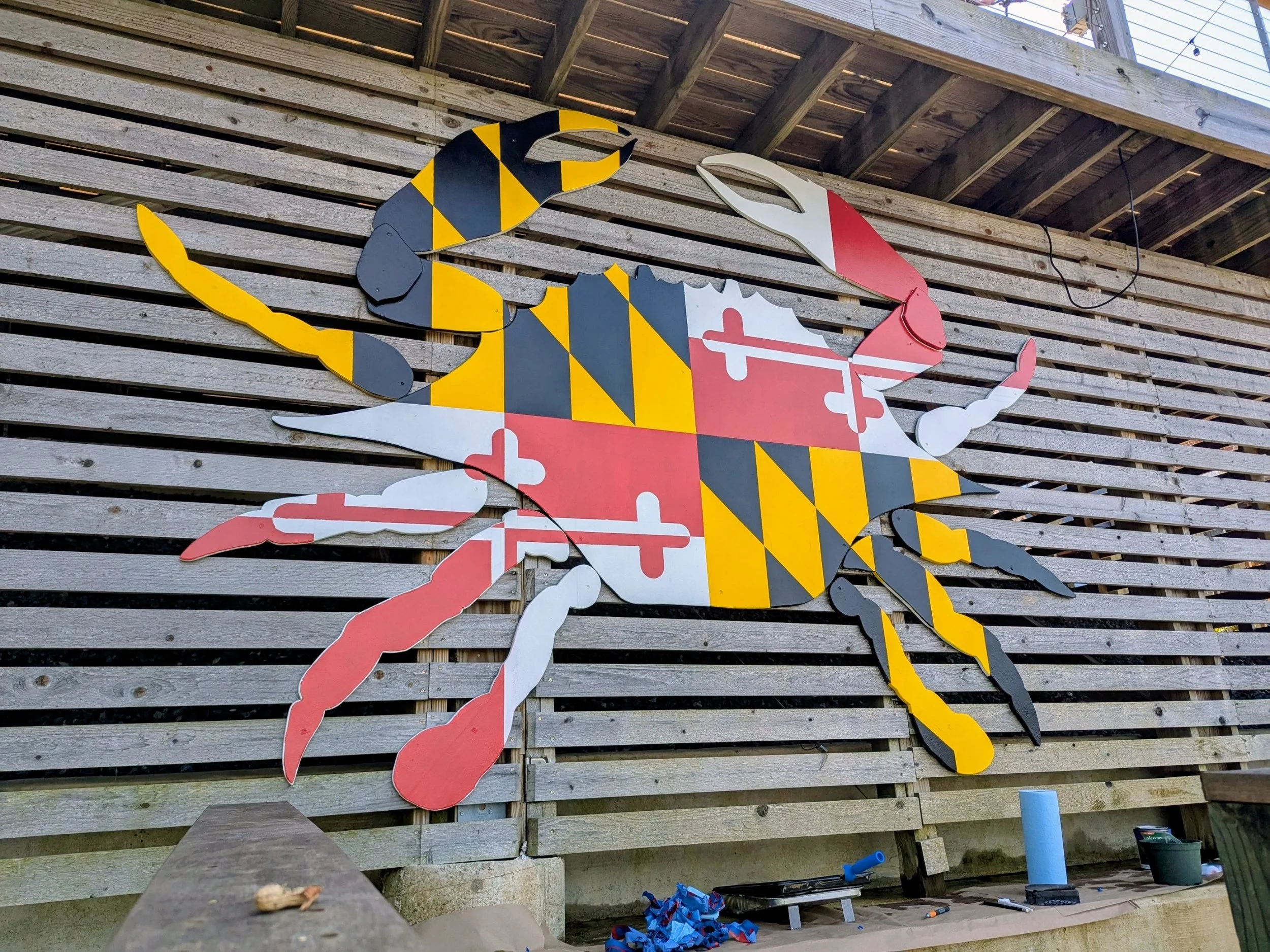A colorful wooden sculpture of a crab with the Maryland state flag design on its body, mounted on a wooden slat wall.