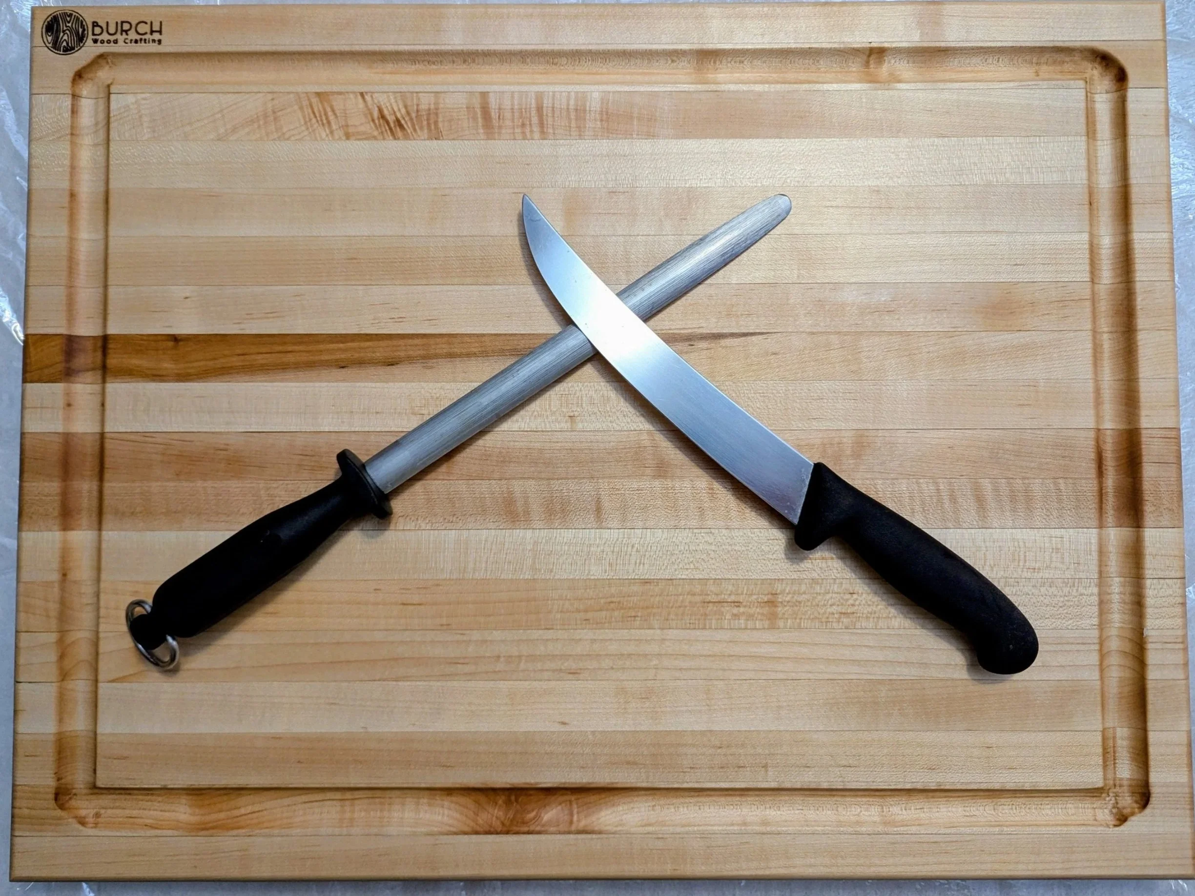 A wooden cutting board with a large kitchen knife and a sharpening steel placed diagonally on it.