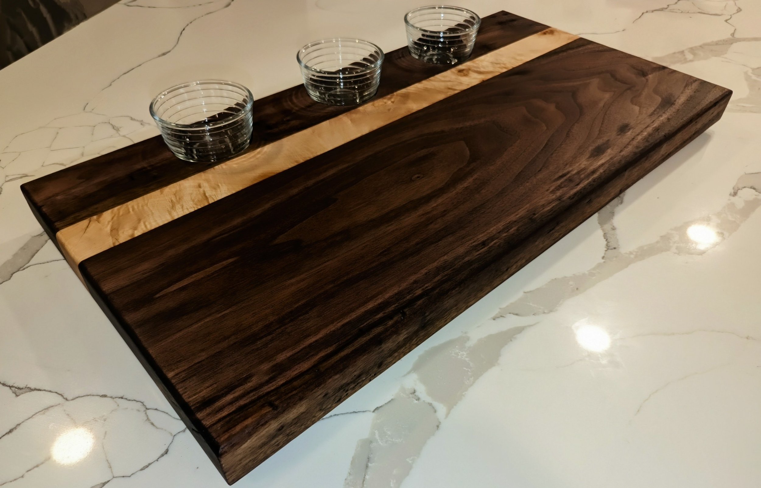 A rectangular wooden serving tray with dark and light wood stripes, placed on a light-colored marble table, with three small glass bowls on it.