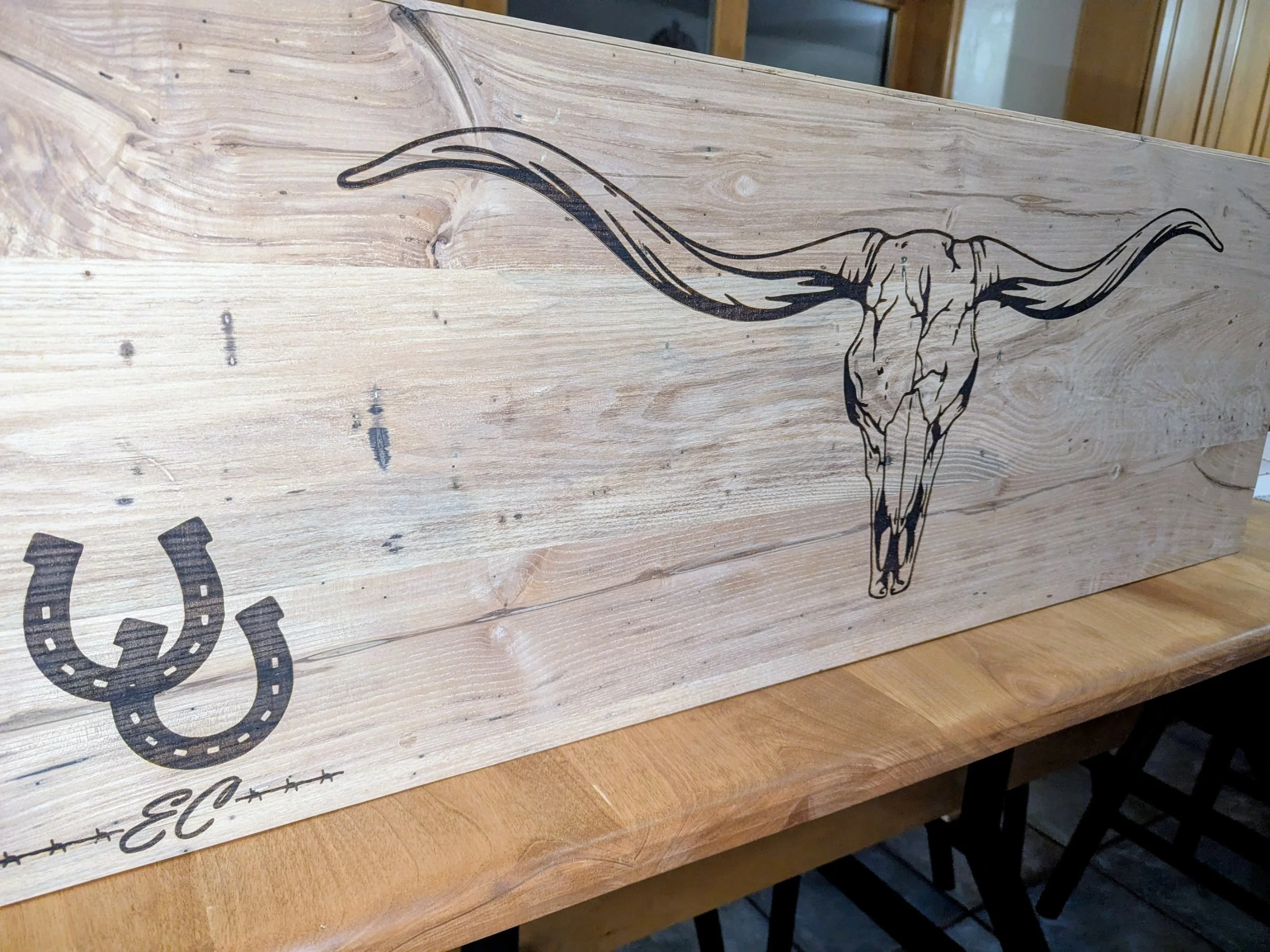 Wooden sign with a black outline illustration of a longhorn skull with large curved horns, and a horseshoe symbol with the name 'El' written below.
