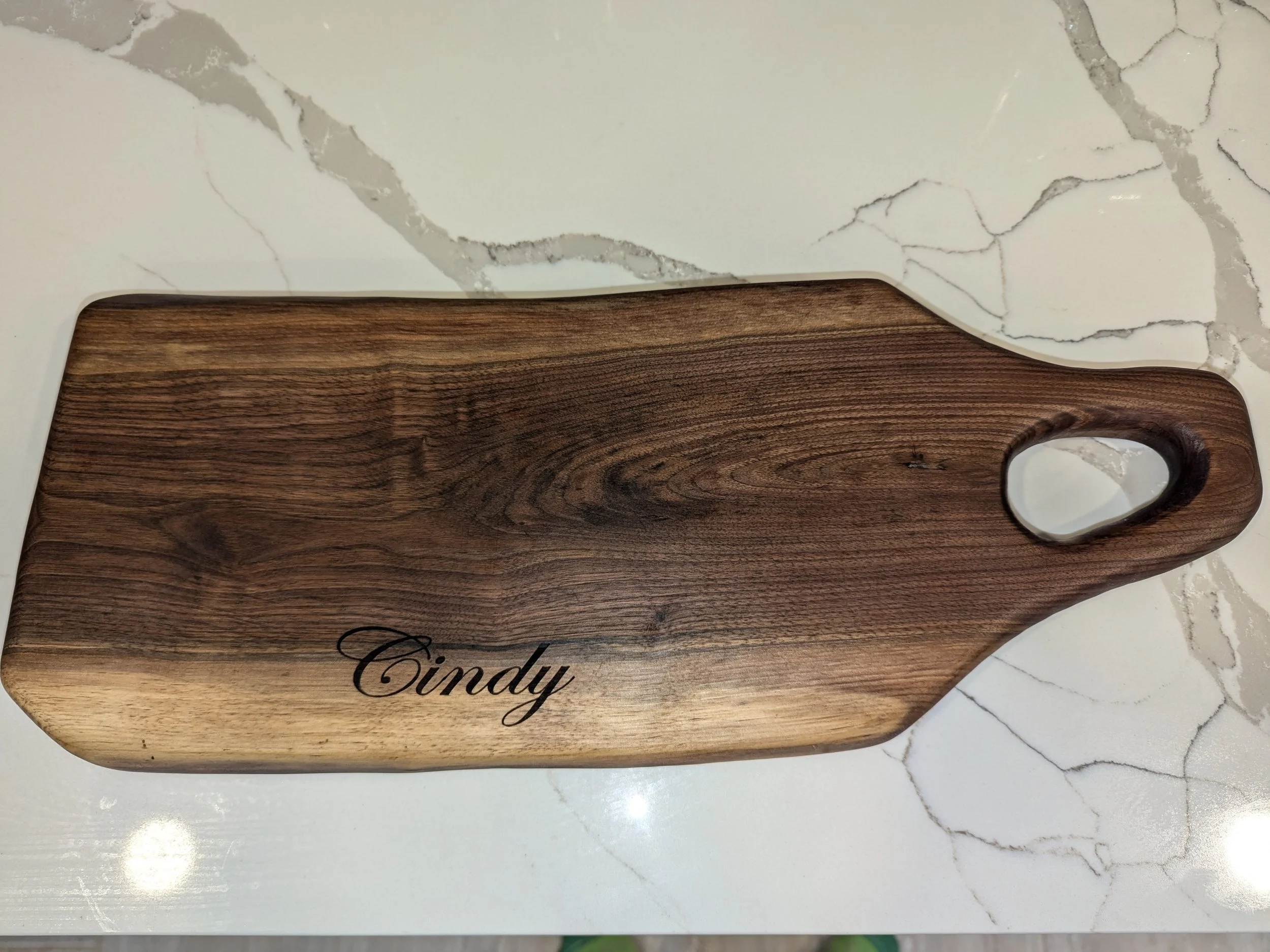 A wooden cutting board with the name 'Cindy' engraved on it, placed on a white marble surface.