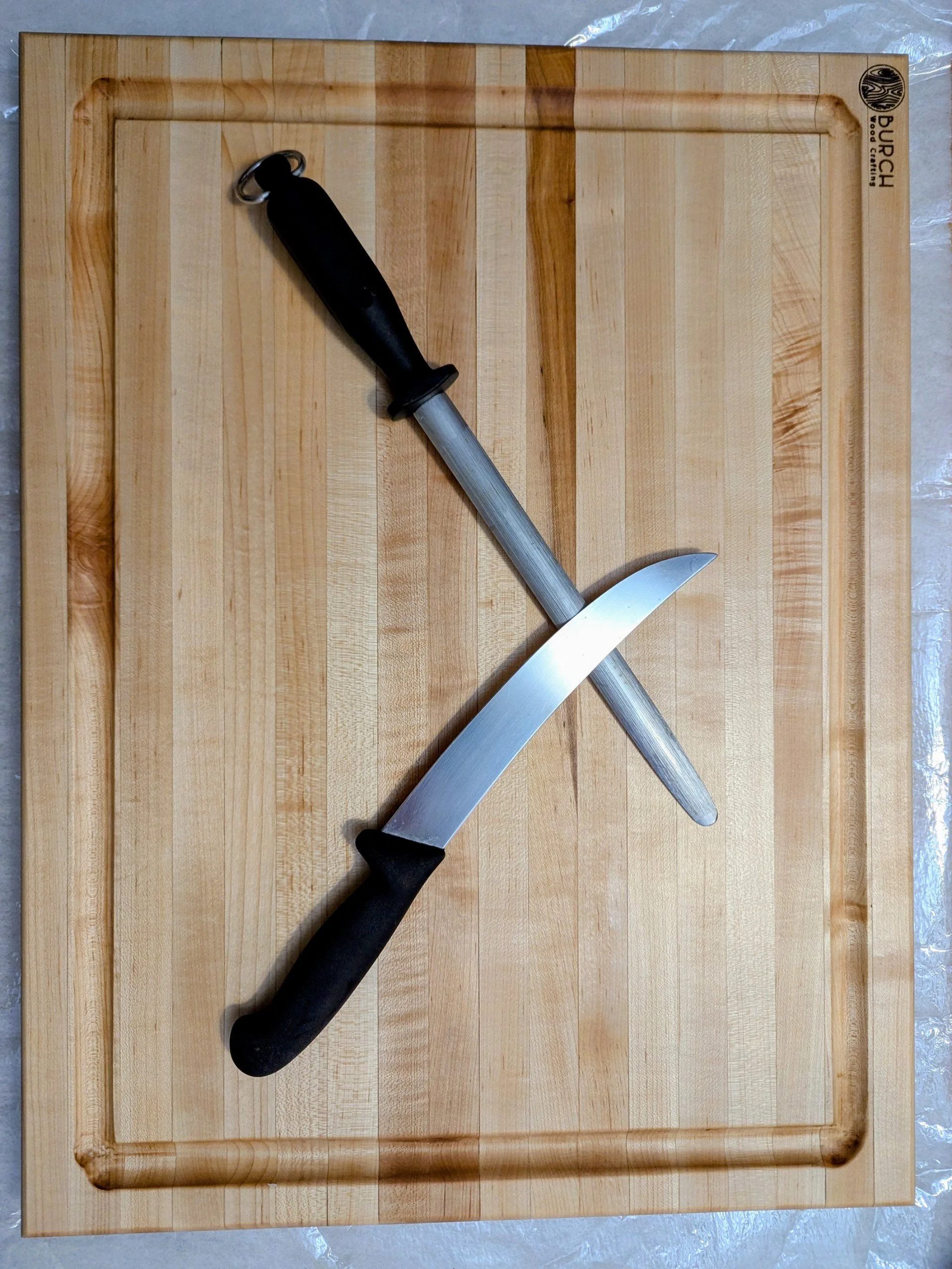 A kitchen cutting board with a black-handled chef's knife and a sharpening steel resting on it.