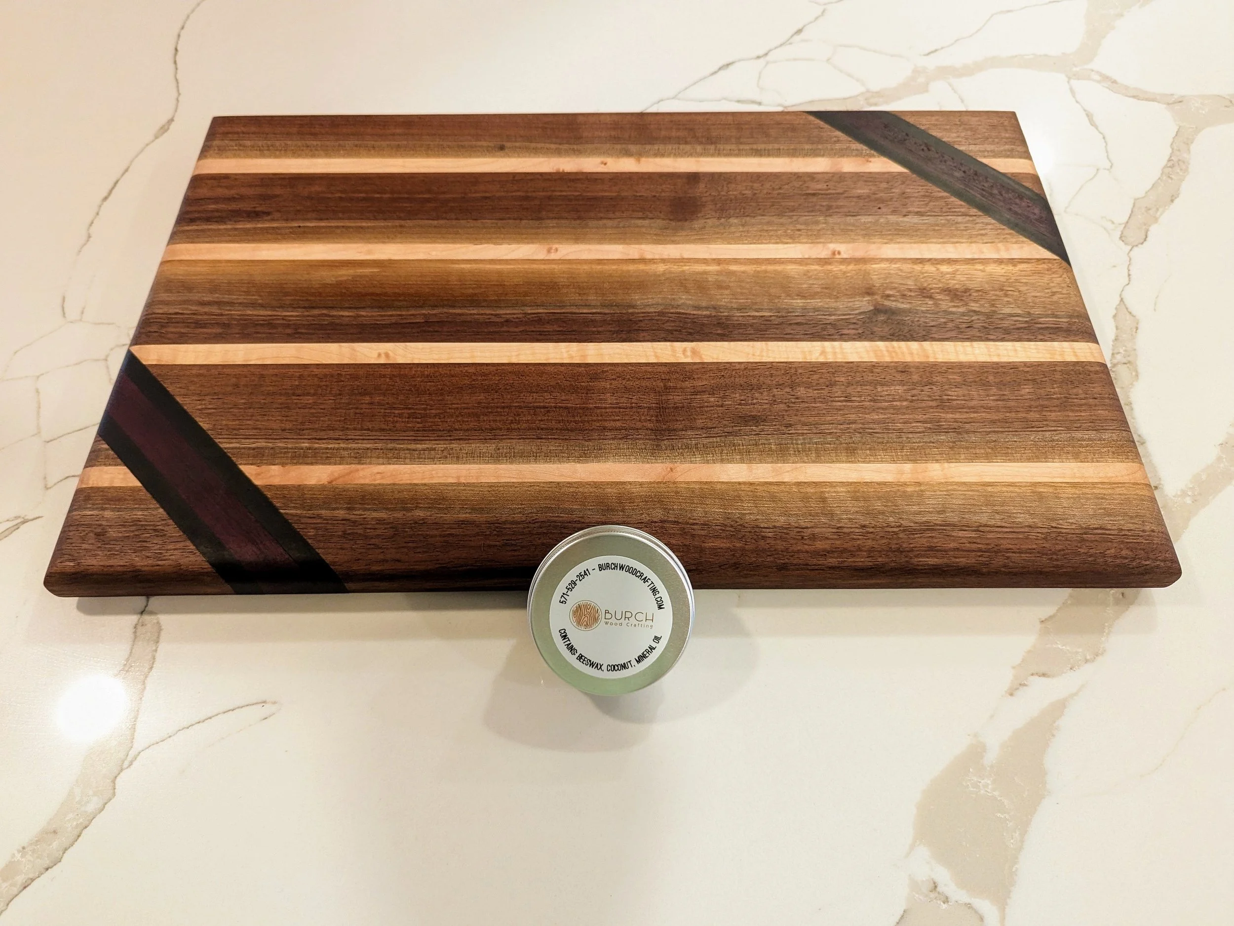 Rectangular wooden cutting board with stripe pattern, placed on a white marble surface with beige and brown veining, and a small round container of wood wax in front of it.