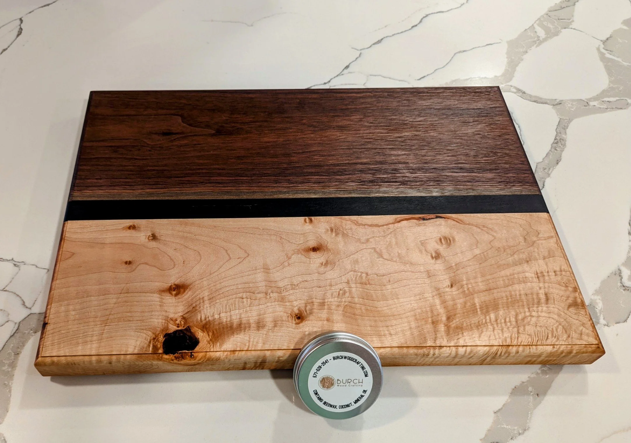 A wooden board with two different types of wood, one dark and one light, separated by a black strip, placed on a white marble surface with gray veins.