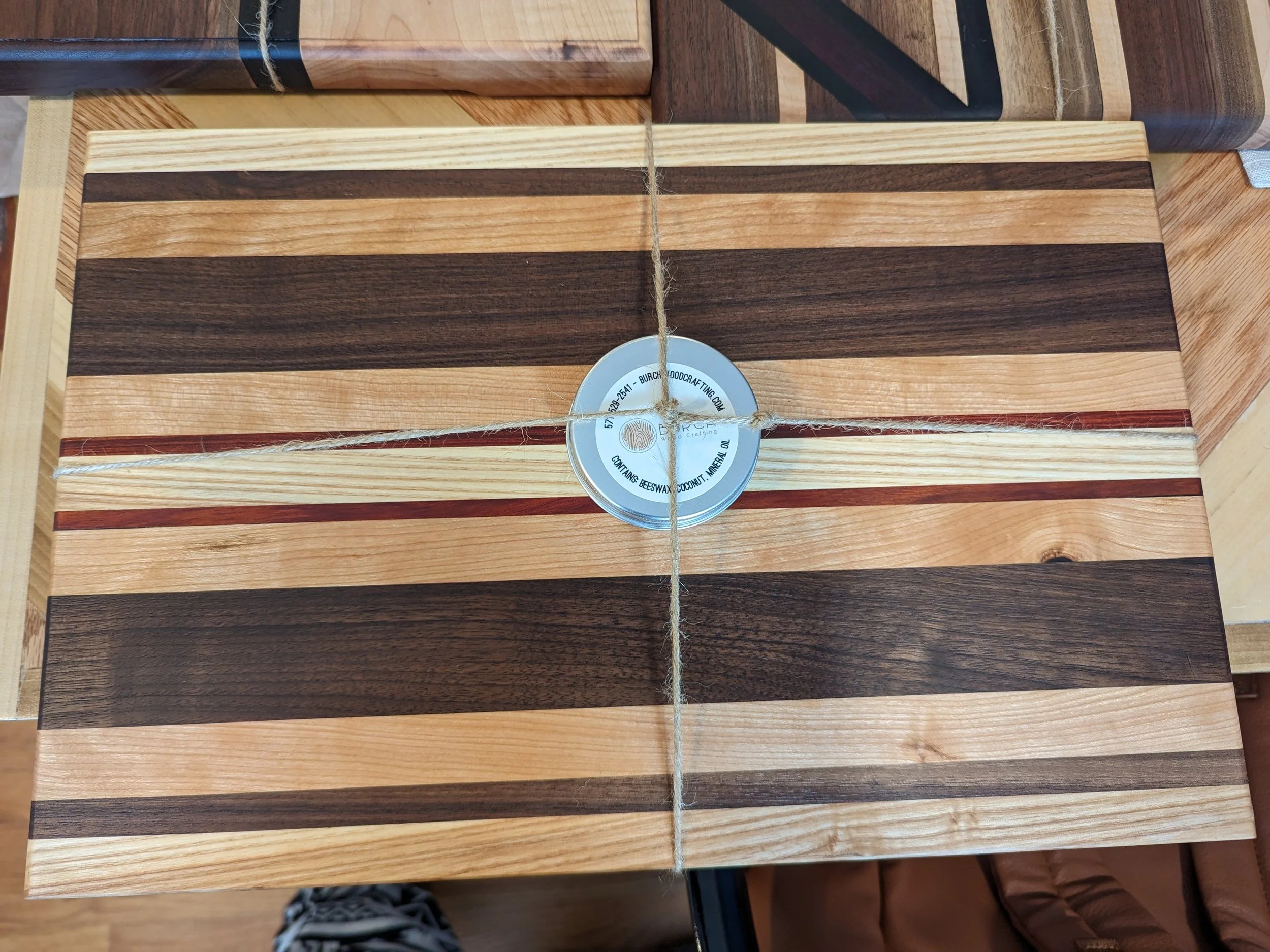 A rectangular wooden cutting board with alternating dark and light wood stripes, wrapped with a piece of twine tied around a tin lid and placed on a table with other wooden surfaces.