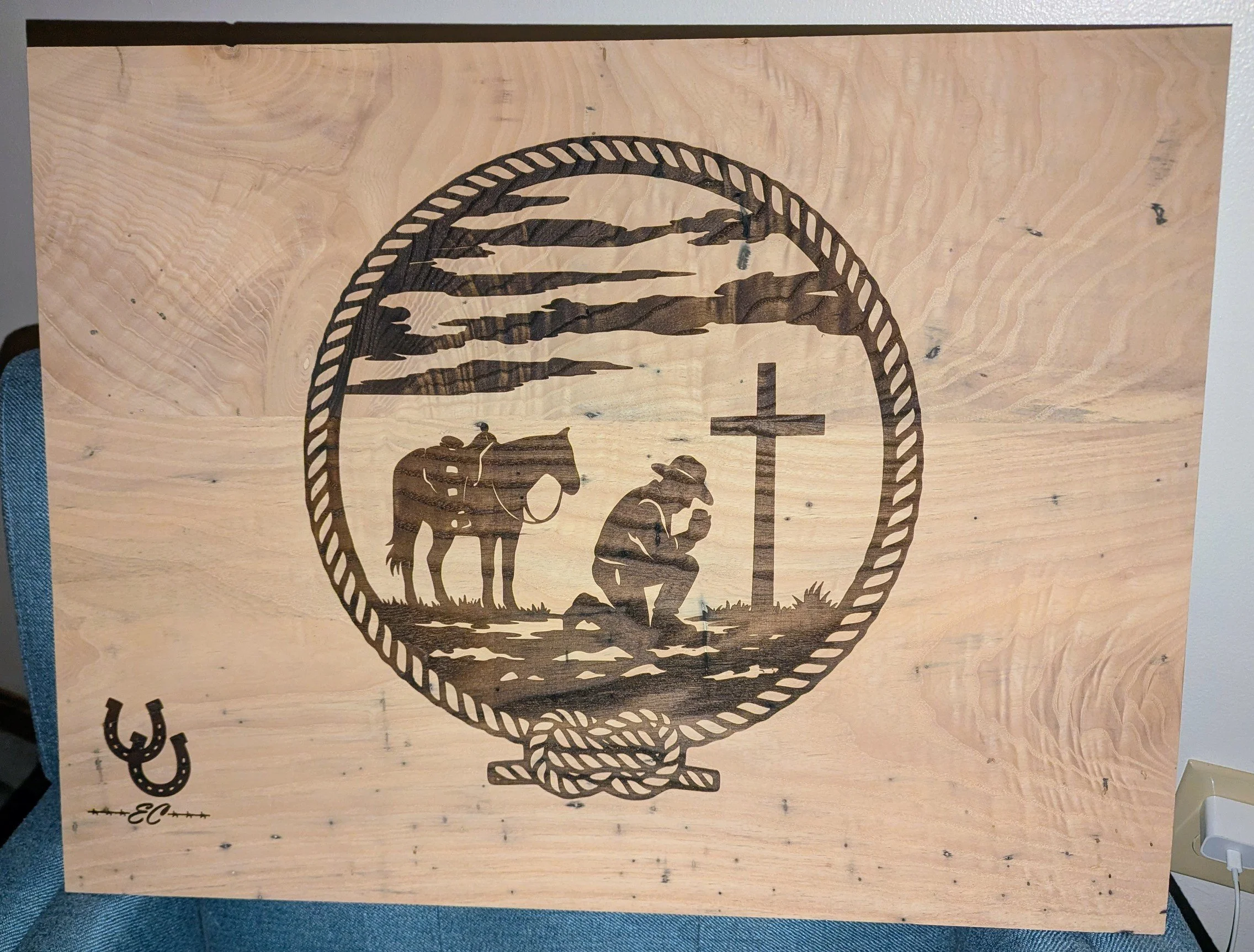 Wooden plaque with a black silhouette of a person kneeling before a cross, with a horse standing nearby, framed by a rope border, and a small horseshoe with initials in the bottom left corner.