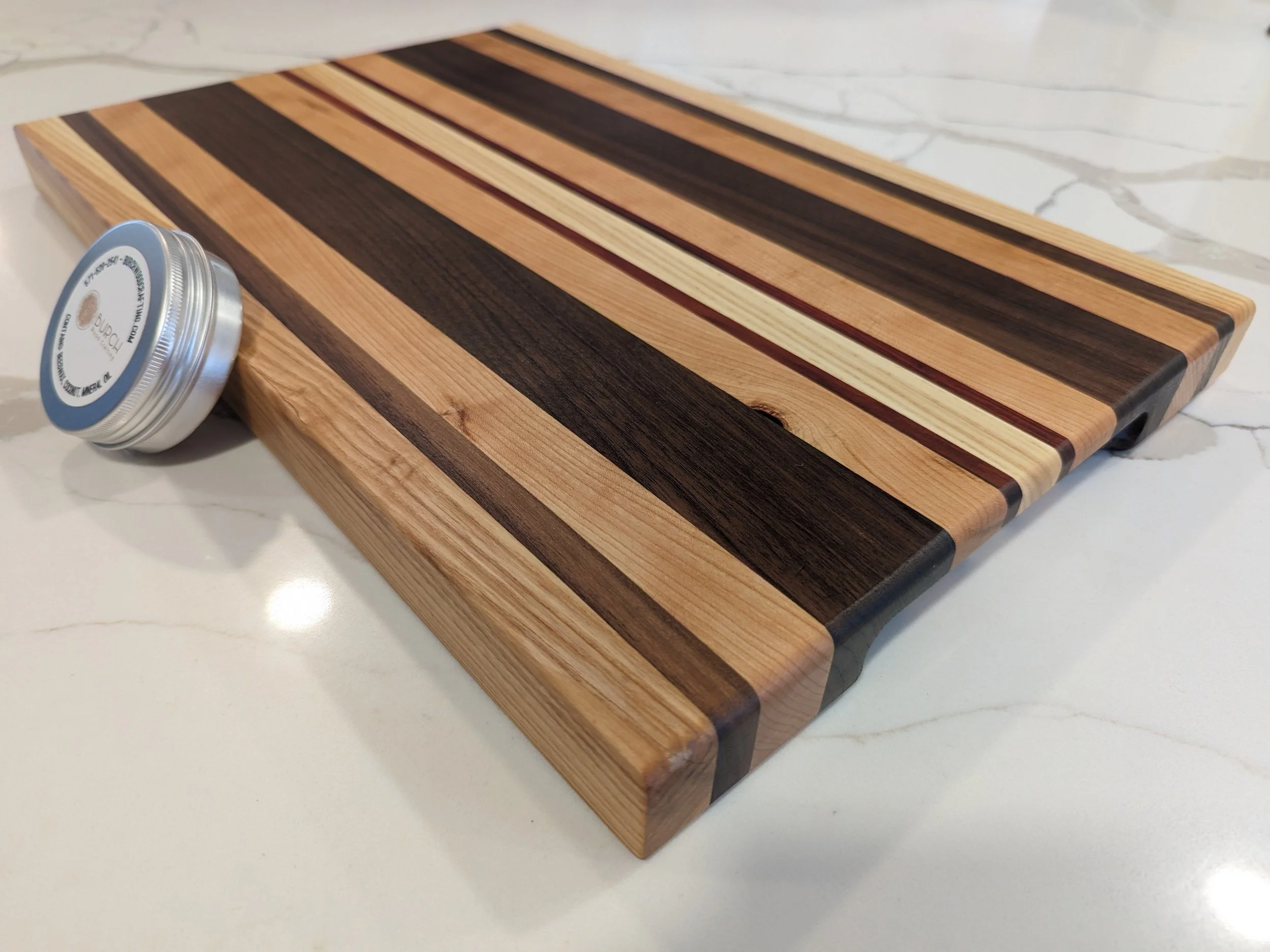 Wooden cutting board with striped pattern of light and dark wood, paired with a small container of seasoning or spice, all placed on a white marble countertop.