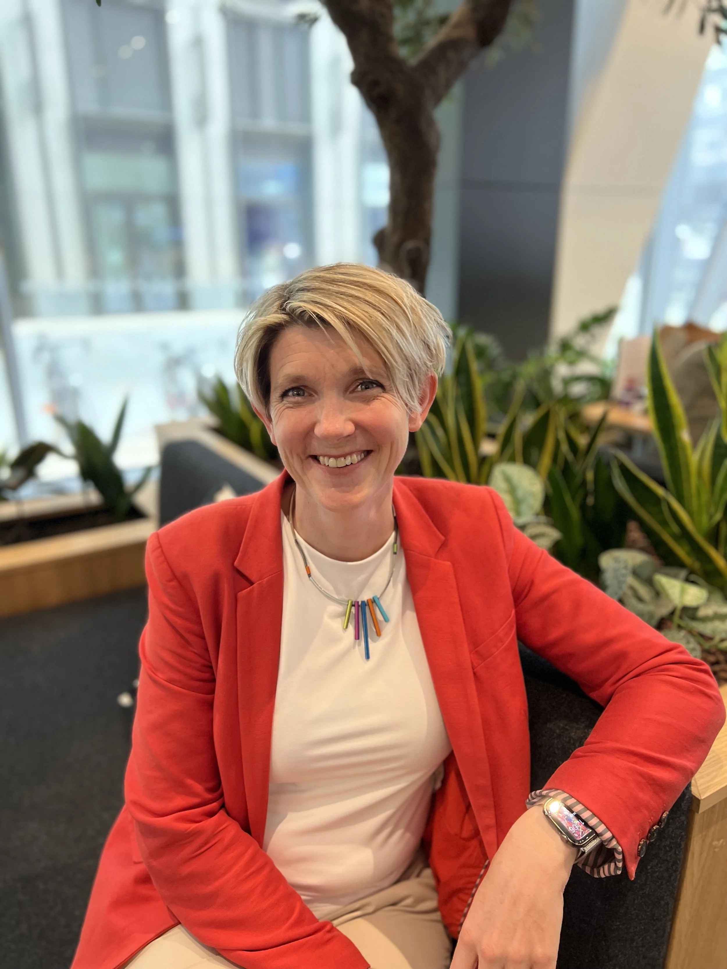 A smiling woman with short blonde hair wearing a red blazer, a white shirt, and a colorful beaded necklace, sitting indoors with large windows and green plants in the background.