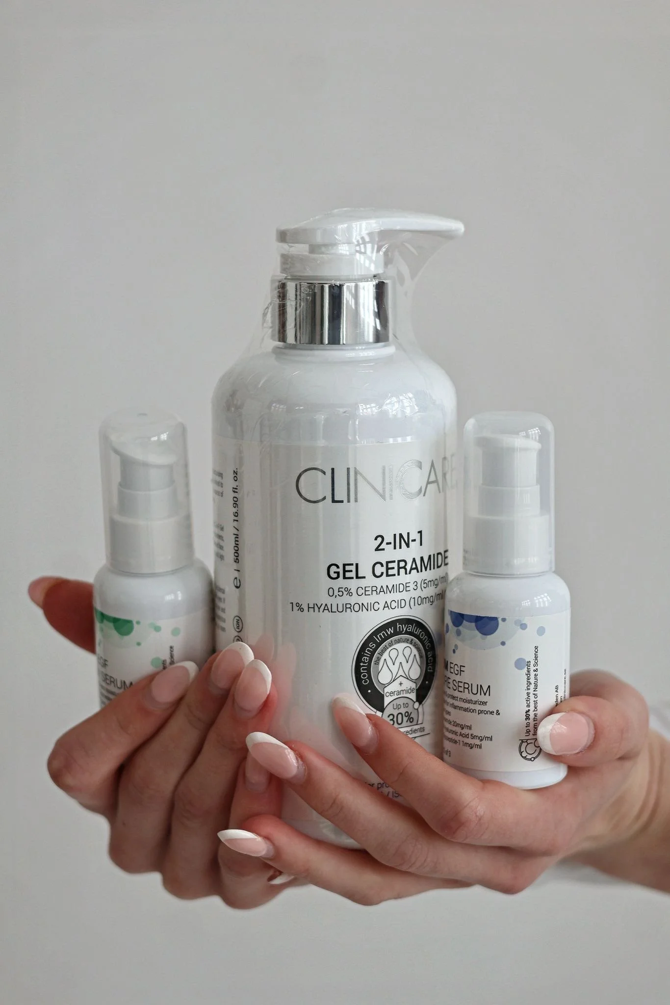 Hands holding three skincare bottles, including a large Clinica 2-in-1 gel with ceramide and hyaluronic acid, and two smaller bottles of serum, against a plain background.