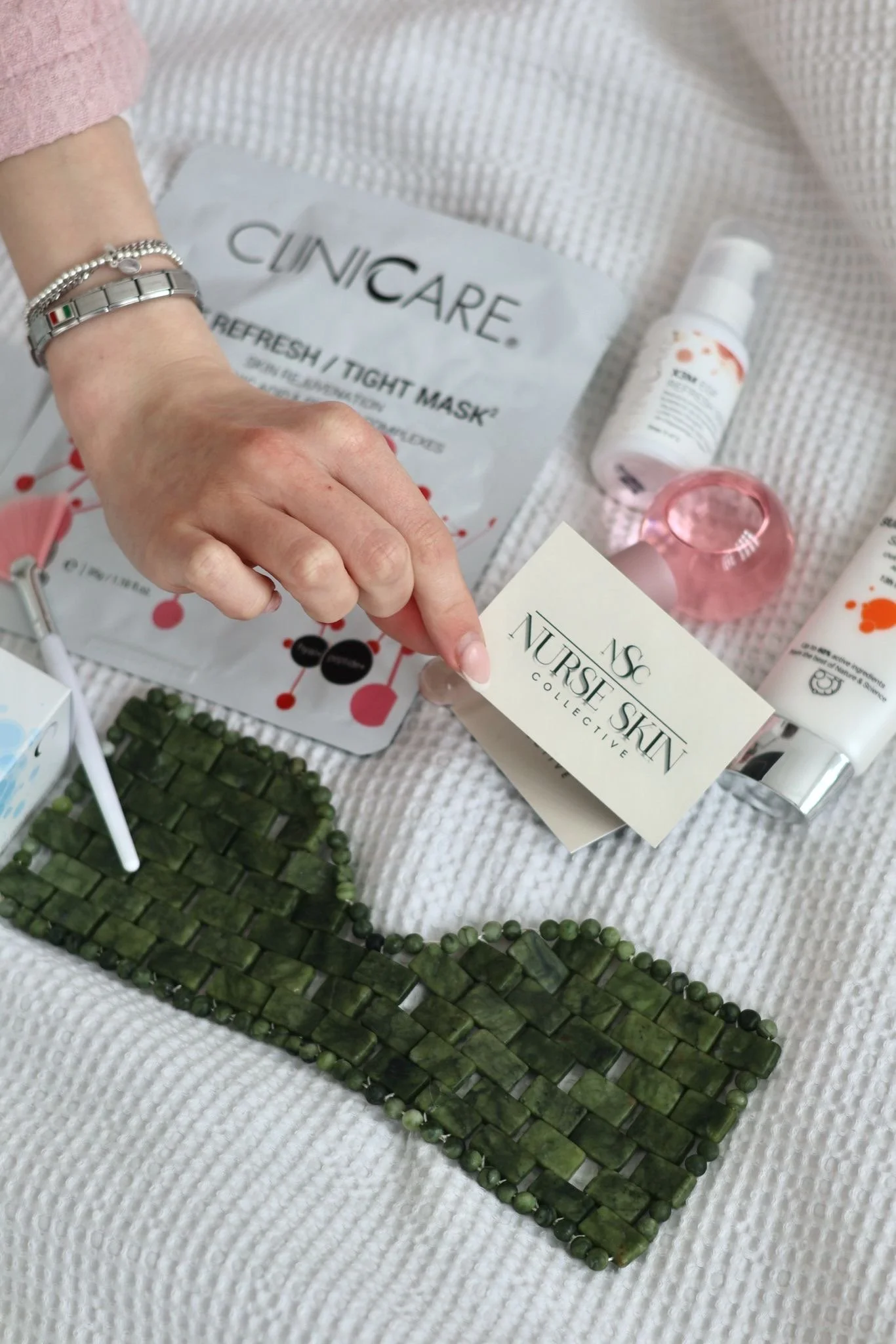 Collection of skincare products with a hand holding a card that reads 'Nurse Skin Collective', a green beaded necklace, and a medical mask on a white textured surface.