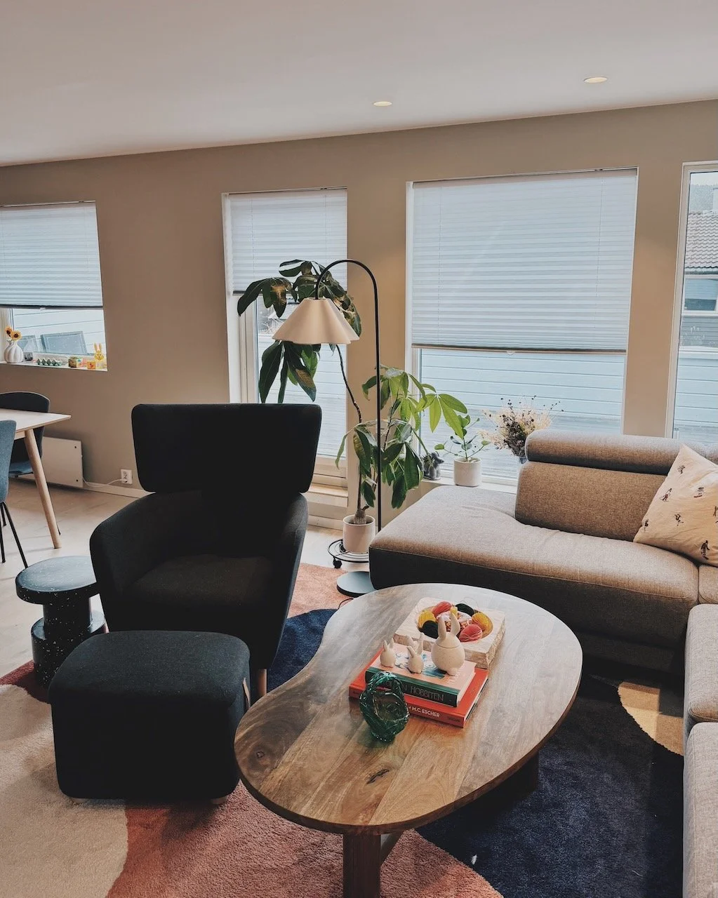 Interiørdesign av Angela Schrøder. Stue med sofa, lenestol, tre vinduer med blinds, planter, salongbord med dekorasjoner.