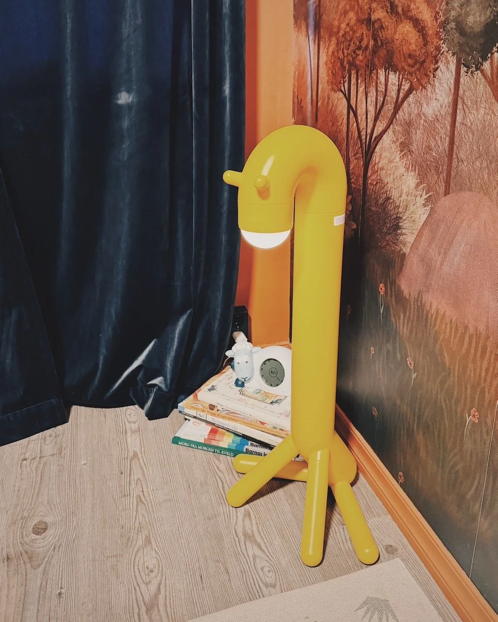 Interiørdesign av Angela Schrøder. Barnerom med girafflampe som står ved en hylle med bøker, omgitt av vegg med naturtapet.