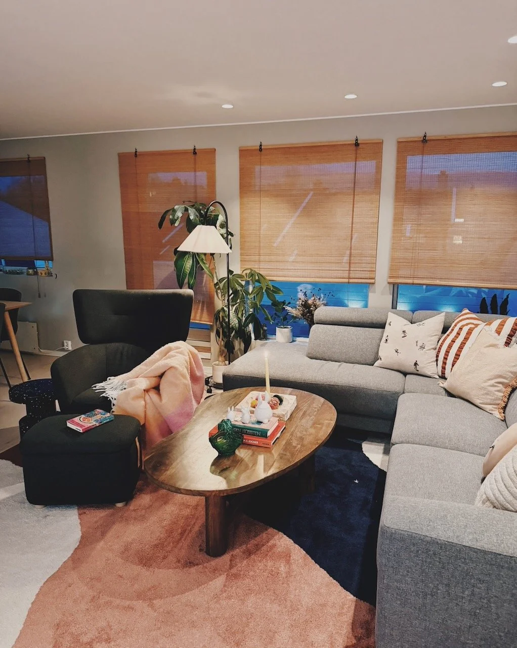 Interiørdesign av Angela Schrøder. Stue med grå hjørnesofa, puffer, blomster, sidebord med bøker og lys, vinduer med rullegardiner, planter, lamper, og puter.