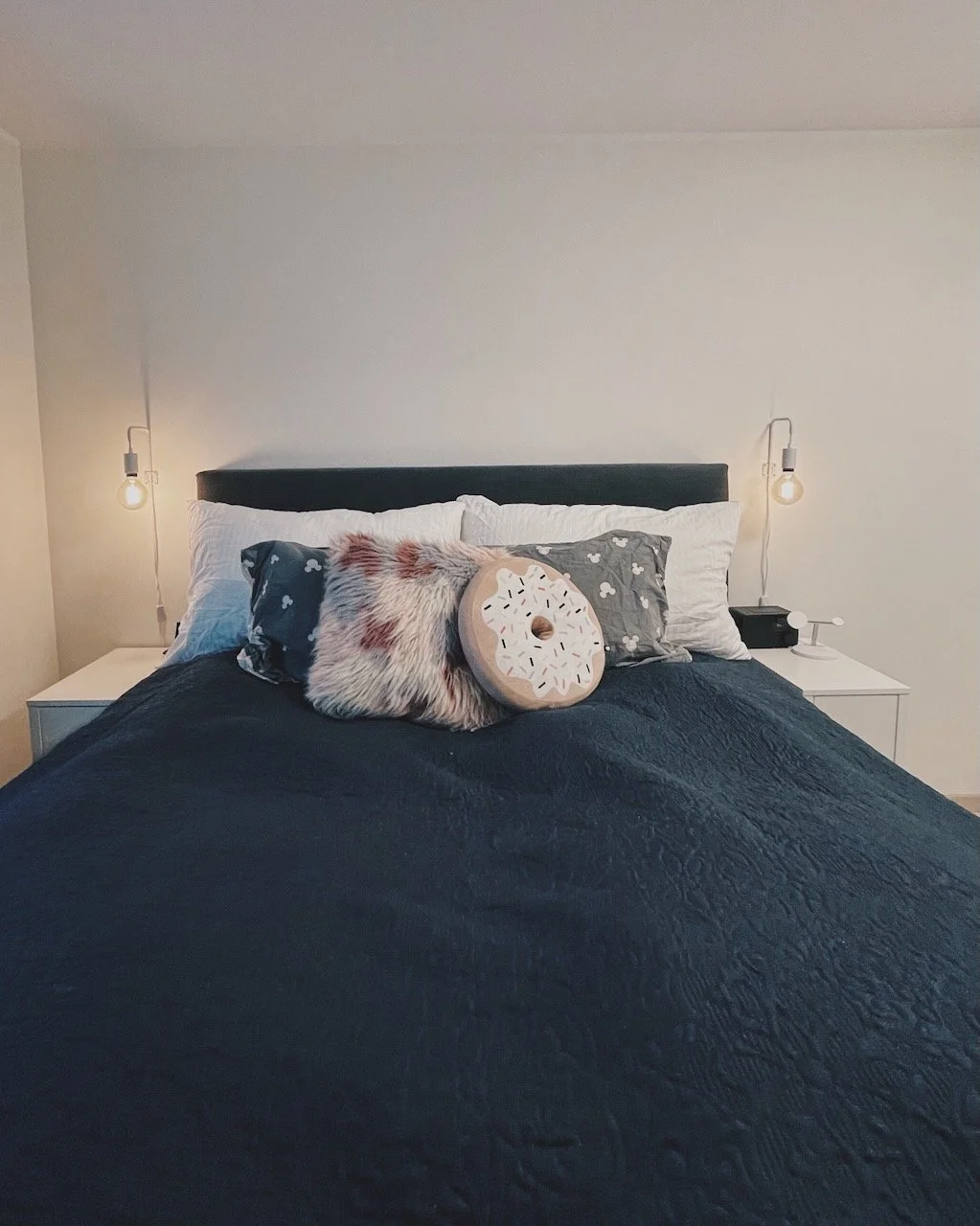 Boligstyling av Angela Schrøder. Seng med flere puter, inkludert en rund pute som ser ut som en donut, og en lampe på hver side av sengen.