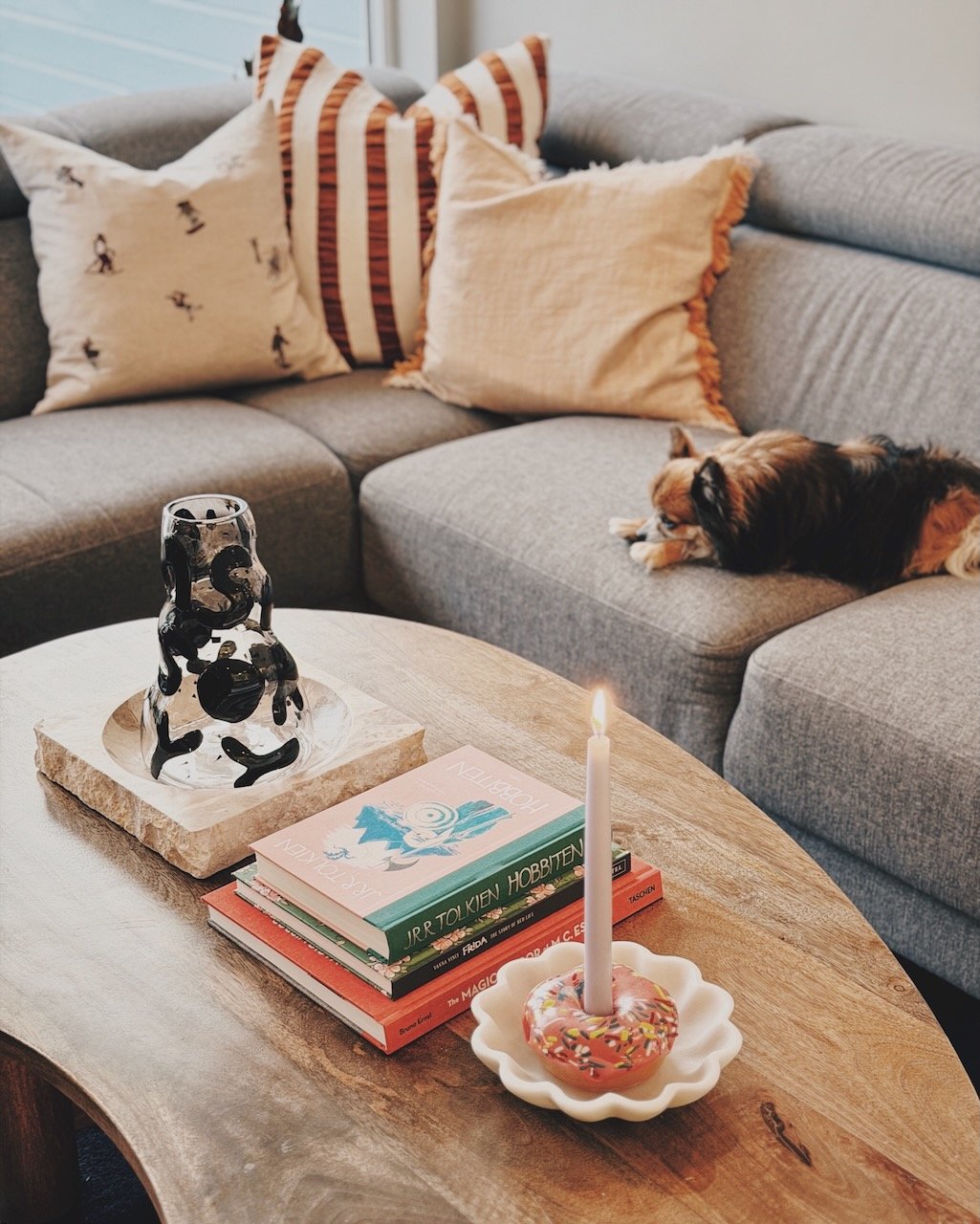 Interiørdesign av Angela Schrøder. En beholder med lys dekorert med konfetti, en stabel bøker, en hvit lysestake med en telys, et grått sofa med flere puter, og en liten hund som ligger på sofaen.