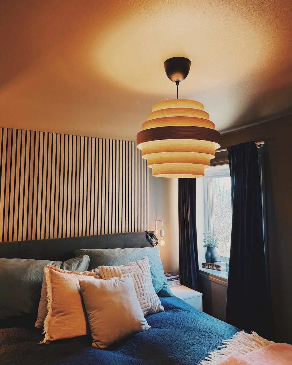 Interiørdesign av Angela Schrøder. Et soverom med en seng dekket av flere puter, et nattbord med bøker, og en stor lysekrone i taket.