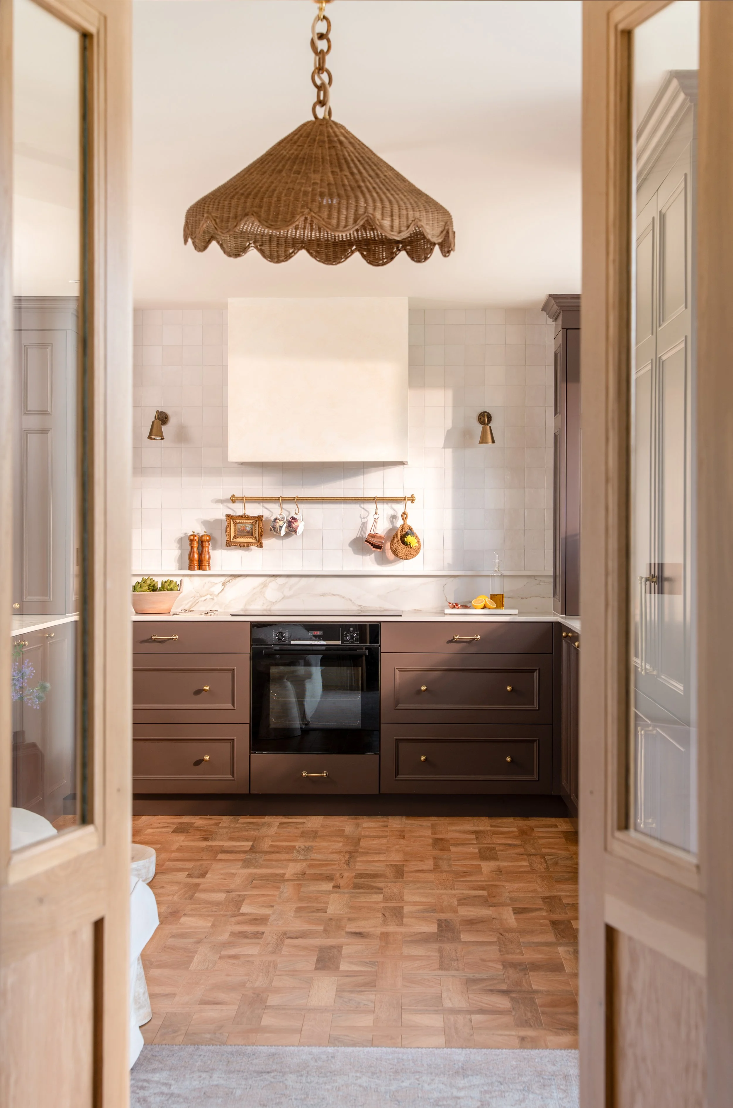View of a kitchen through open wooden doors with glass panels, featuring dark brown cabinets, a black oven, and a white tiled wall with hanging kitchen utensils and decorations. Wooden flooring and a beige rug are also visible.