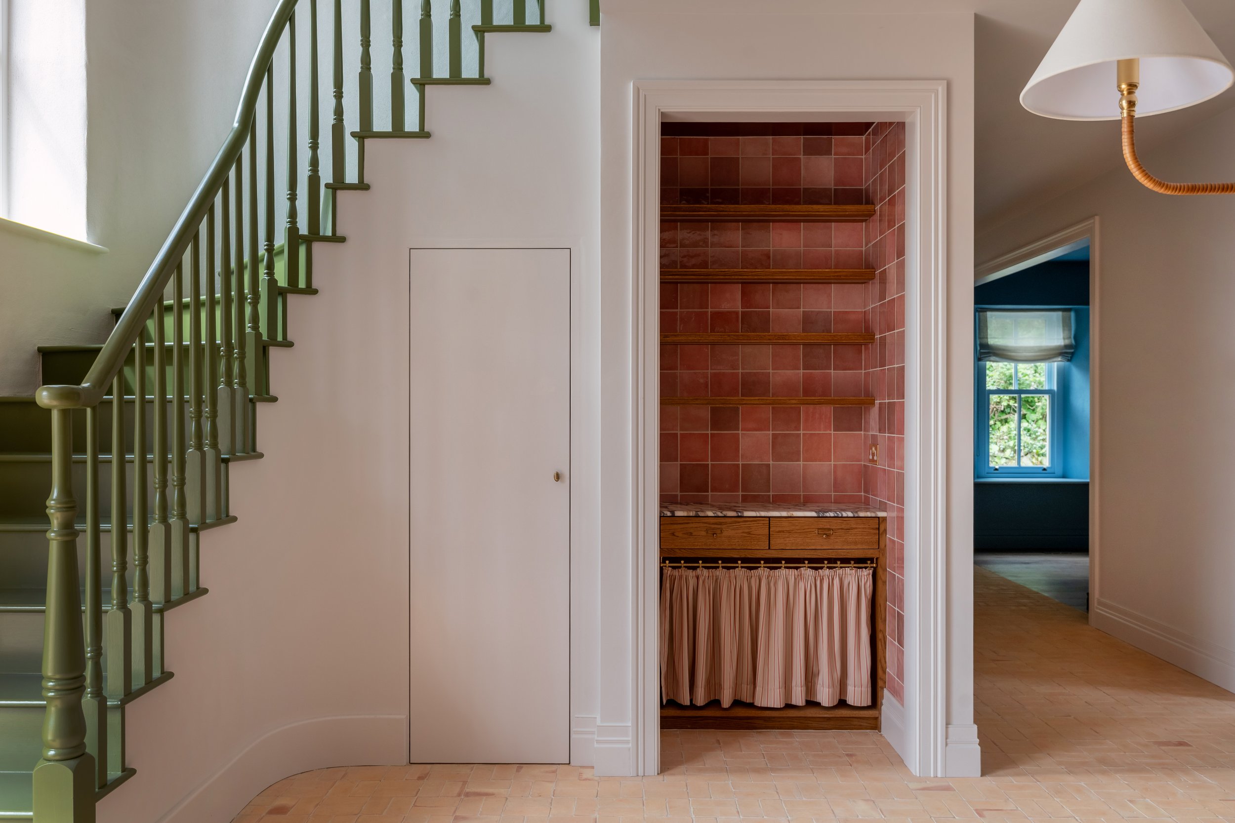 Completed hallway at Deerpark Residence, Co. Clare, by Aoife Maria Interiors, featuring an olive-green staircase and a bespoke oak storage alcove with terracotta tiles and pink fabric.