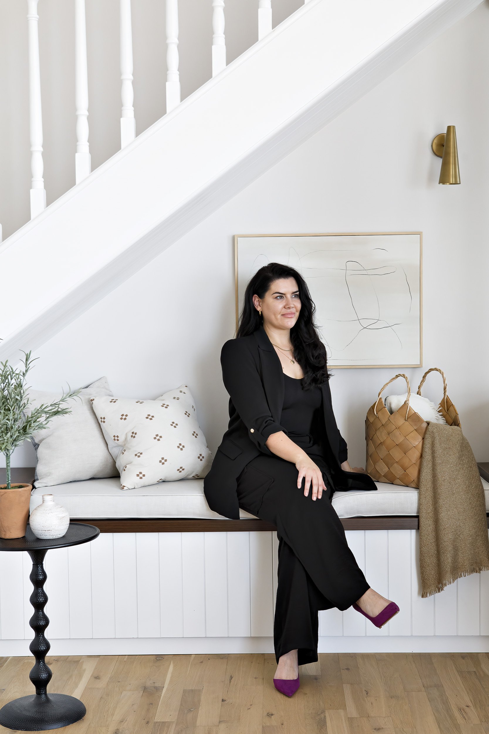 A woman with long dark hair sitting on a built-in white bench under a staircase, wearing a black blazer and matching pants with purple heels. The area is decorated with pillows, a small potted plant, a white vase on a black side table, and a large woven bag. There is abstract art on the wall and a gold wall sconce.