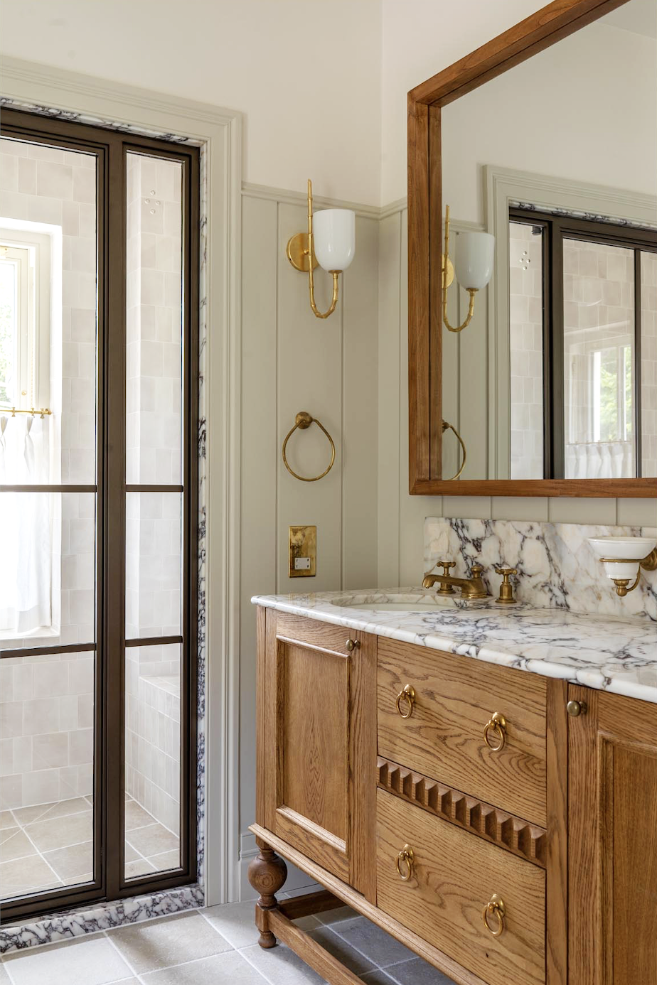 A bathroom vanity with a marble countertop, wooden cabinet, and a large wall-mounted mirror with a wooden frame. Wall-mounted light fixtures with gold accents, a brass towel ring, and a soap dish are also visible. A glass shower door is seen on the left.
