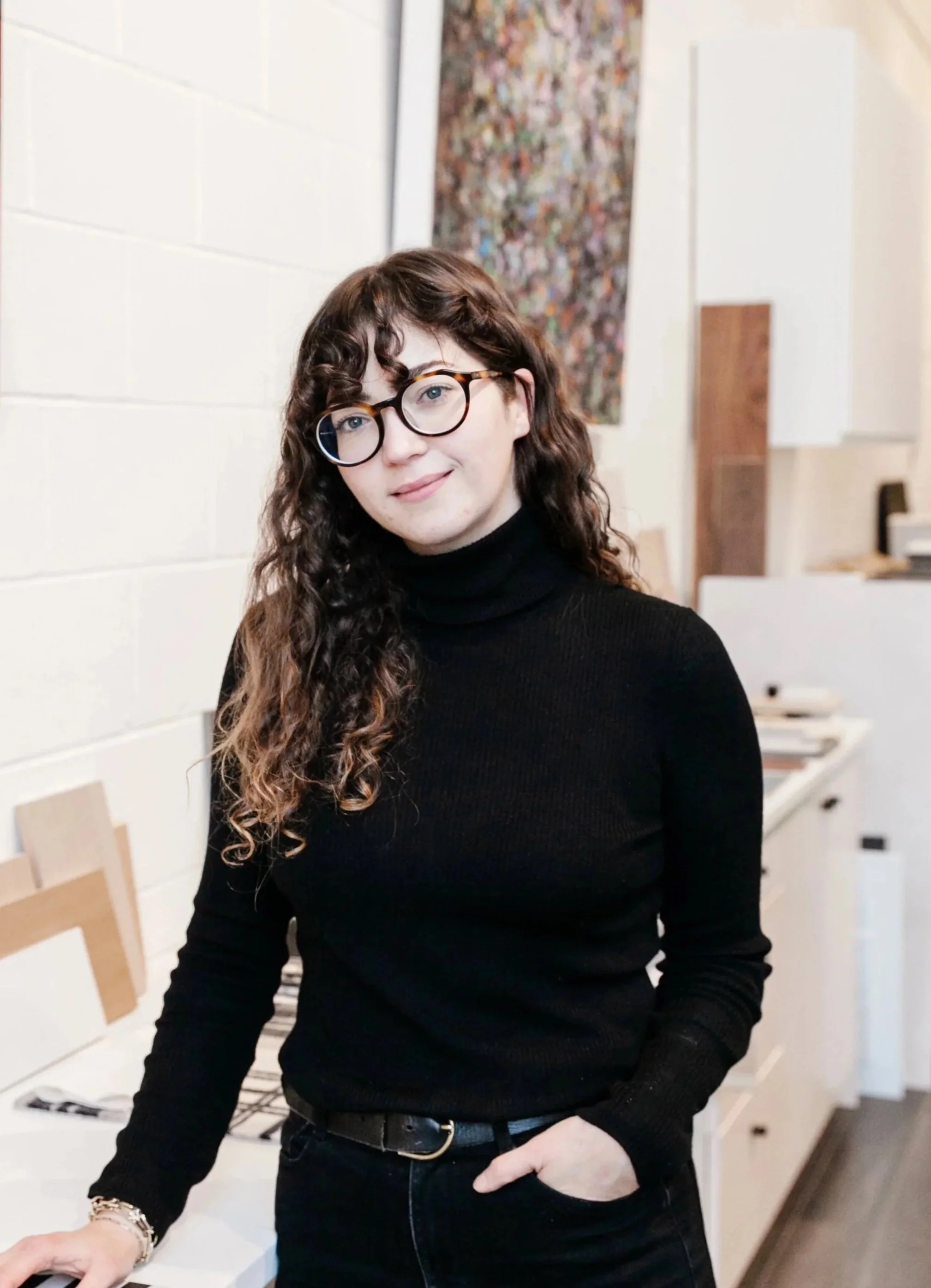 A woman with curly brown hair and glasses, wearing a black turtleneck, standing in an art studio or workspace with white walls and artworks in the background.