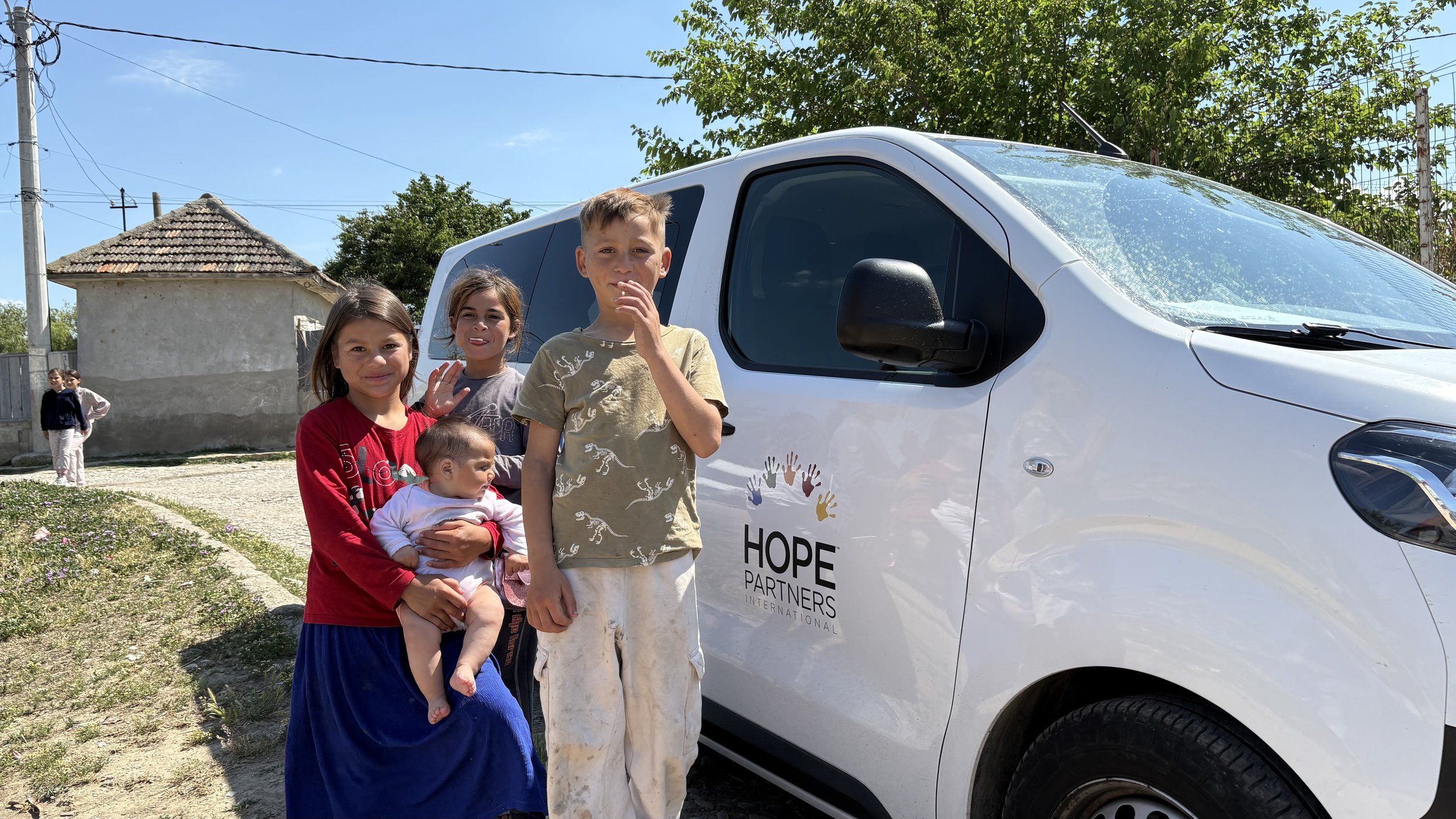 Four children stand in front of a white van with the logo 'Hope Partners International' on the side. A small girl is being held by a girl in a red long-sleeve shirt and blue skirt. Next to them, a boy in a beige shirt with animal prints and white pan