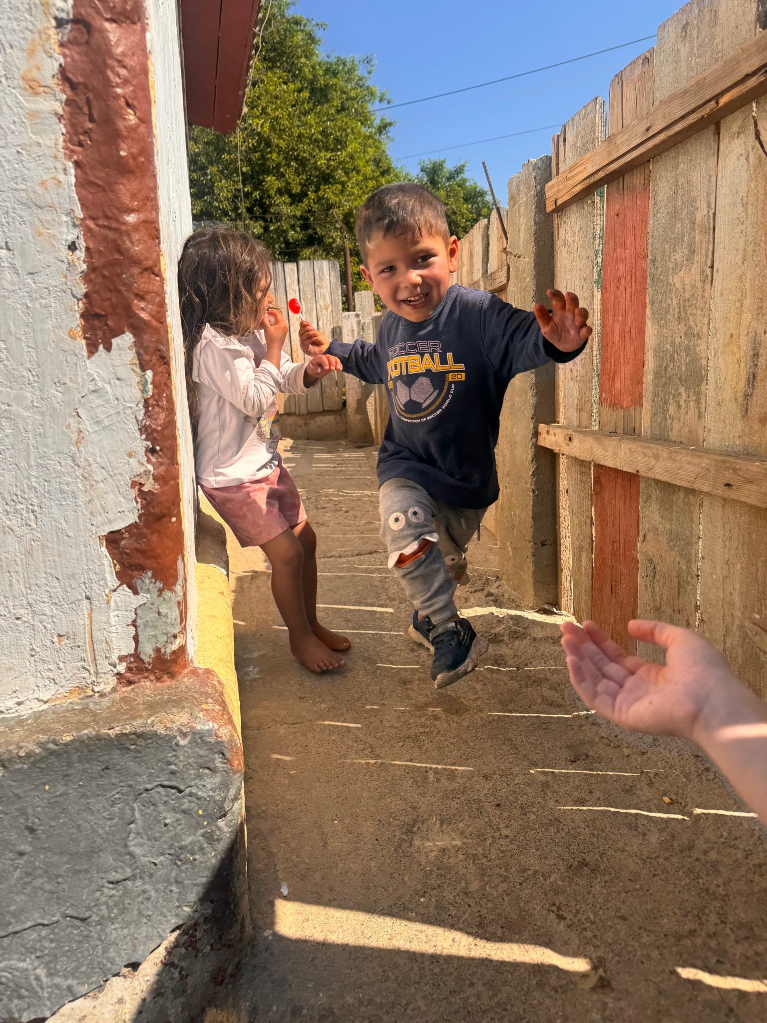 A boy jumping in the air and two children sitting behind him in a narrow outdoor alley between a white building and a wooden fence.