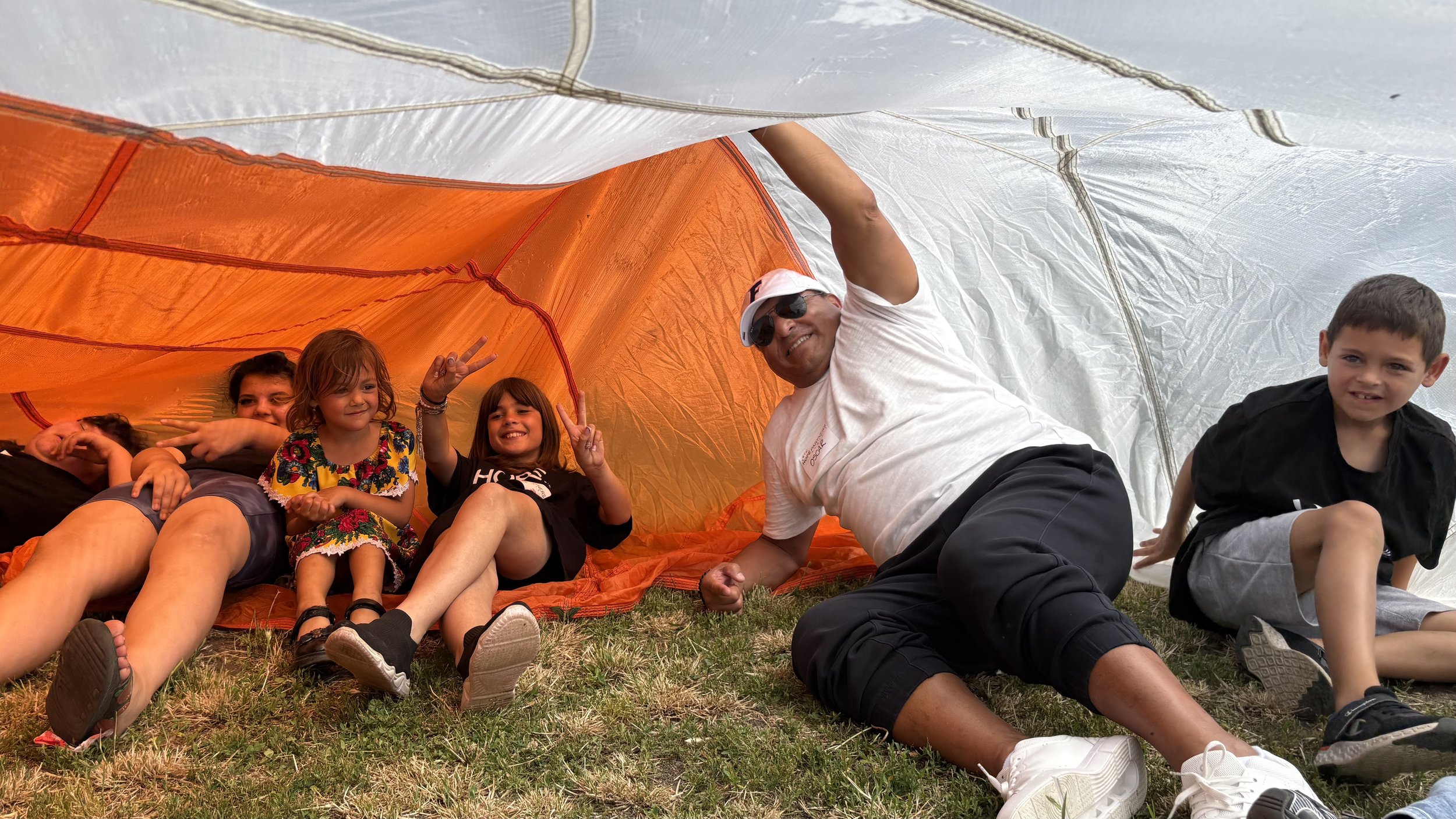 Children and an adult inside a camping tent, sitting on grass, smiling and making peace signs, with the tent's orange interior and white exterior visible.
