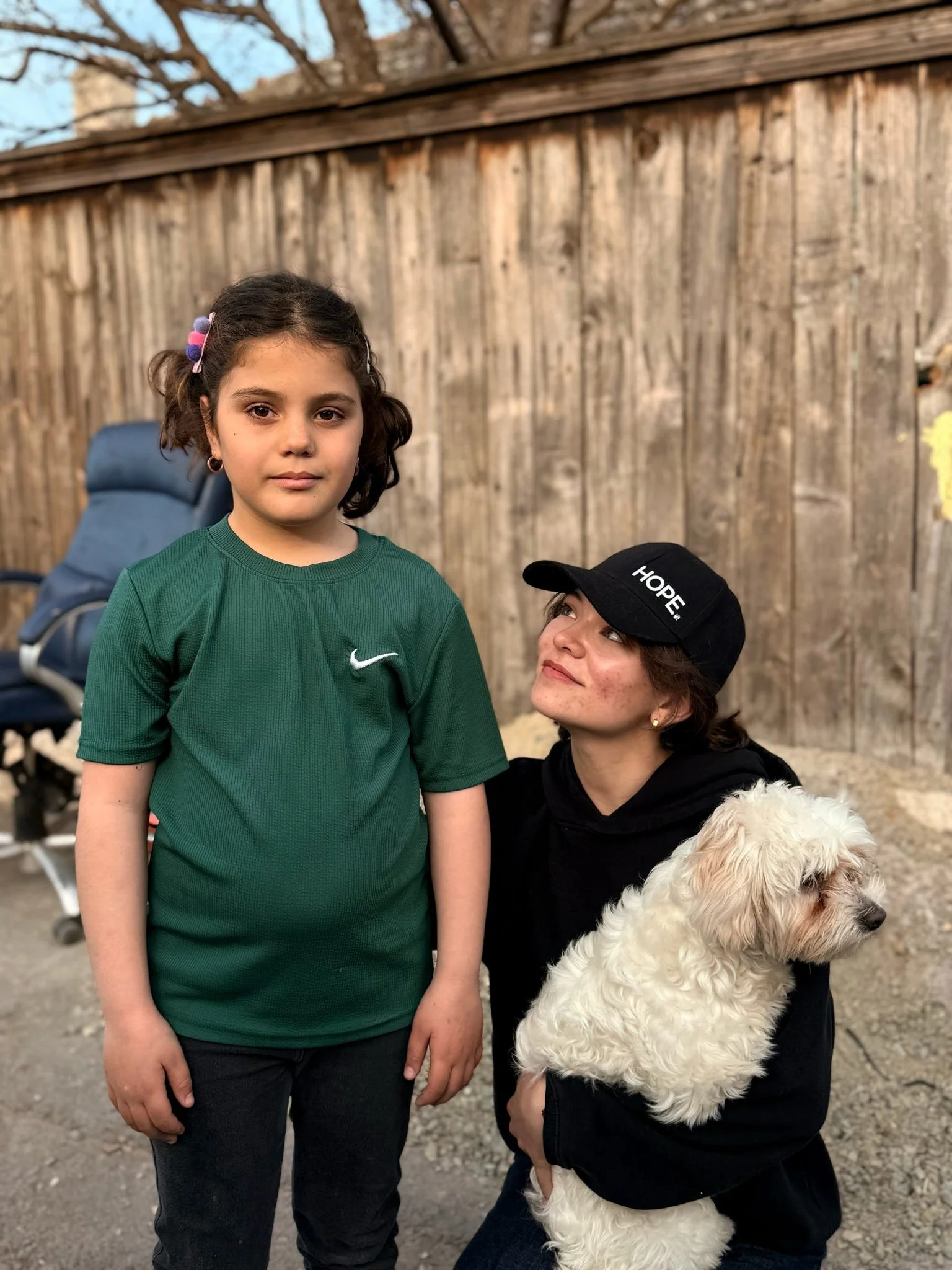 A young girl with brown hair and a green shirt standing outdoors next to a woman with short hair, a black cap with 'HOPE' written on it, and a black hoodie. The woman is holding a fluffy white dog. They are in front of a wooden fence.