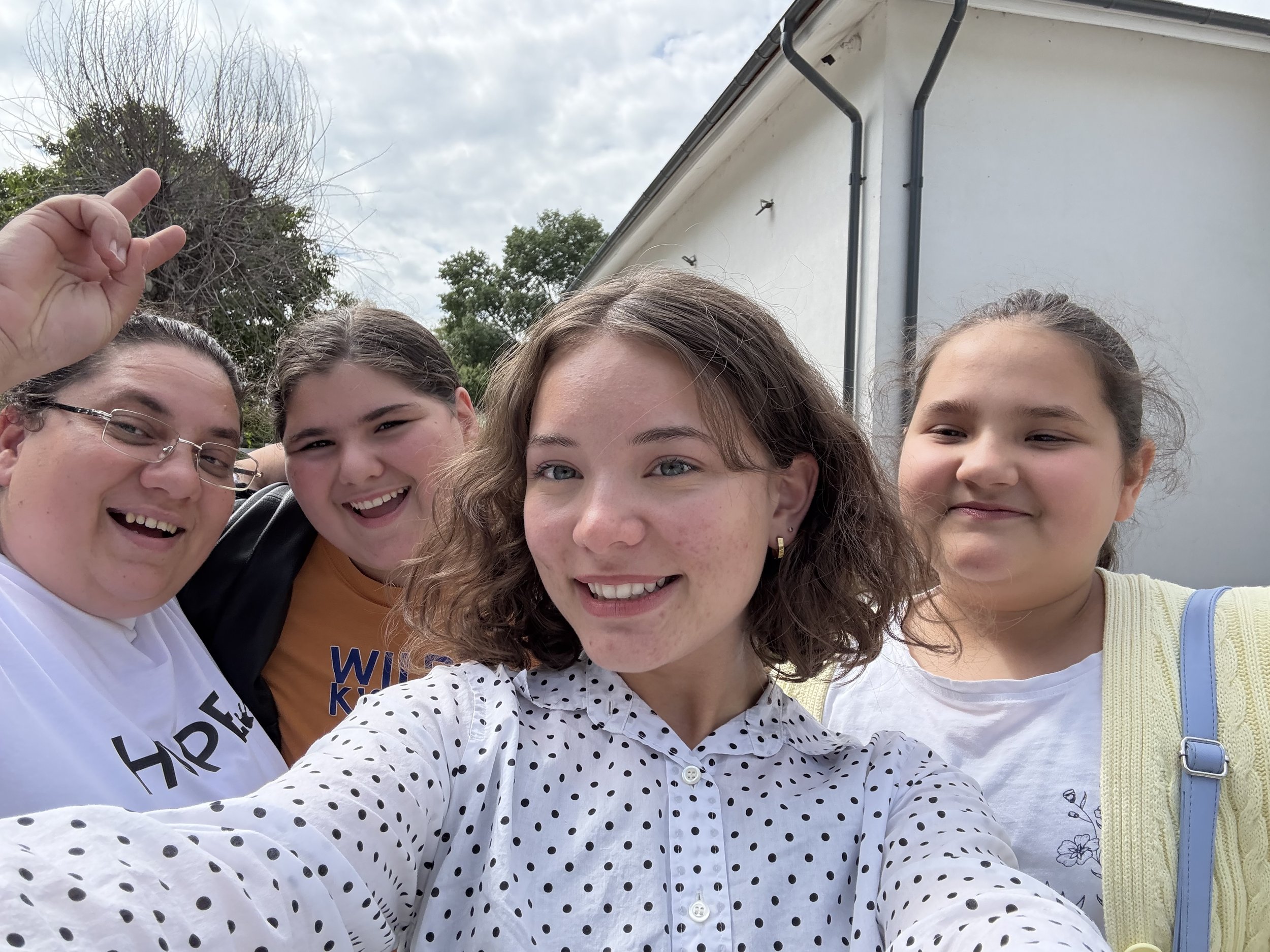 Group of five young women smiling and taking a selfie outdoors, with trees and a building in the background.