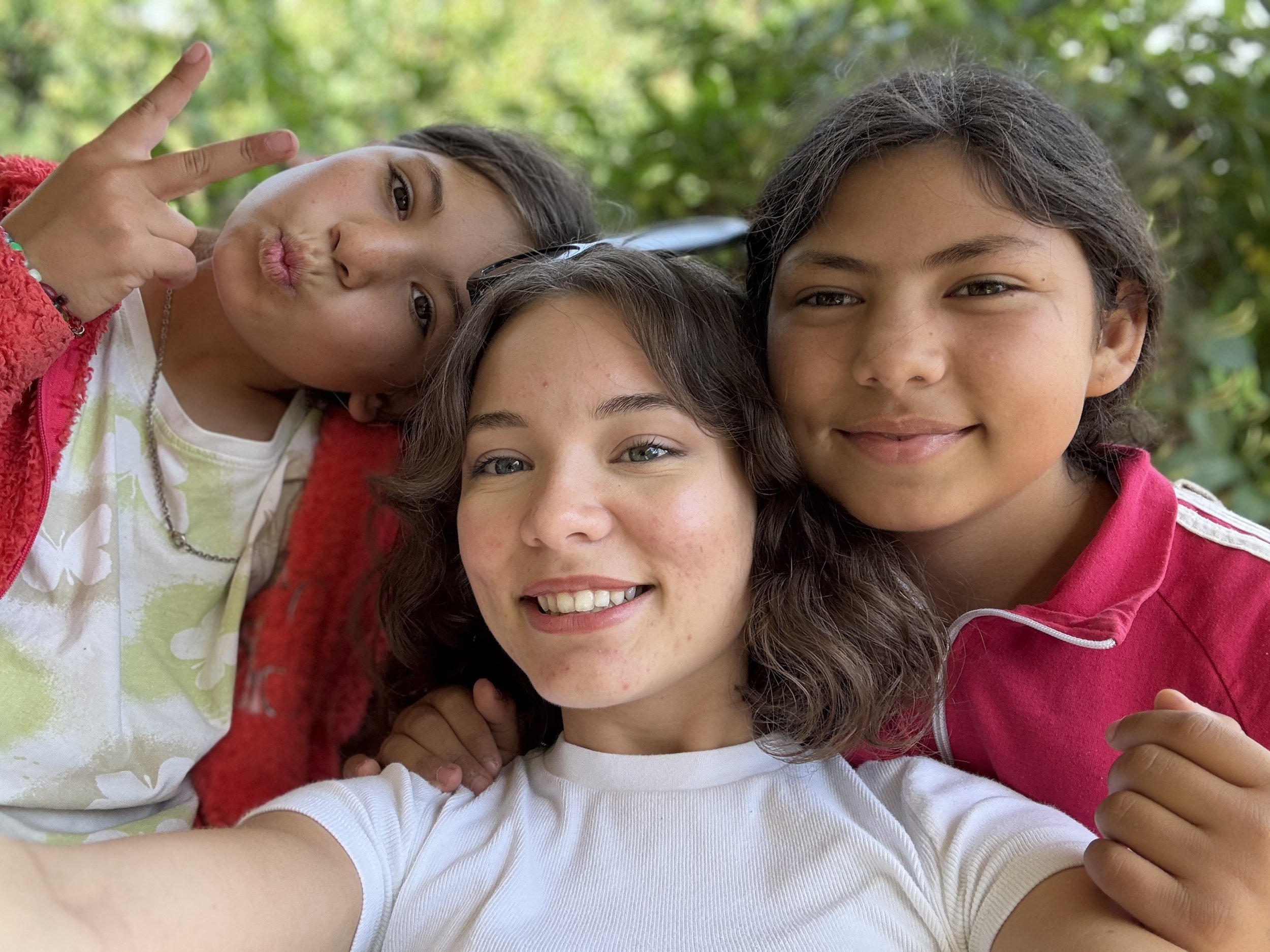 Three girls taking a selfie outdoors with green foliage in the background.