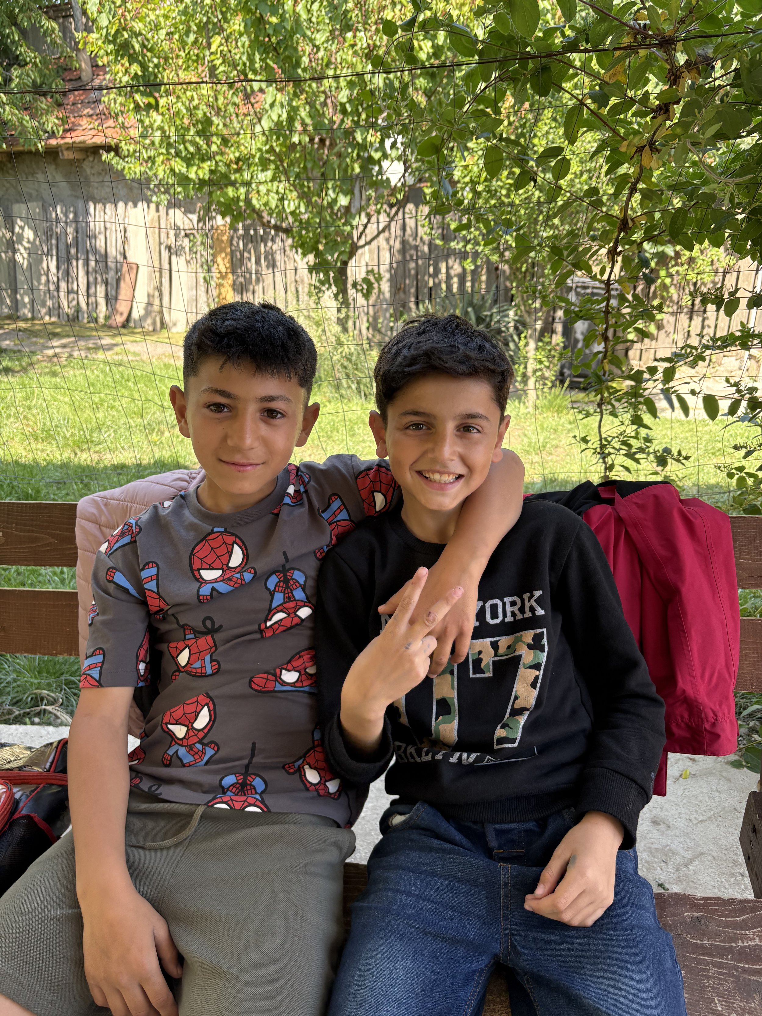 Two boys sitting on a park bench outdoors, smiling, with one arm around each other's shoulders. One boy is wearing a Spider-Man patterned T-shirt, and the other is wearing a black hoodie with a large '7' on it. A pink backpack is on the bench next to