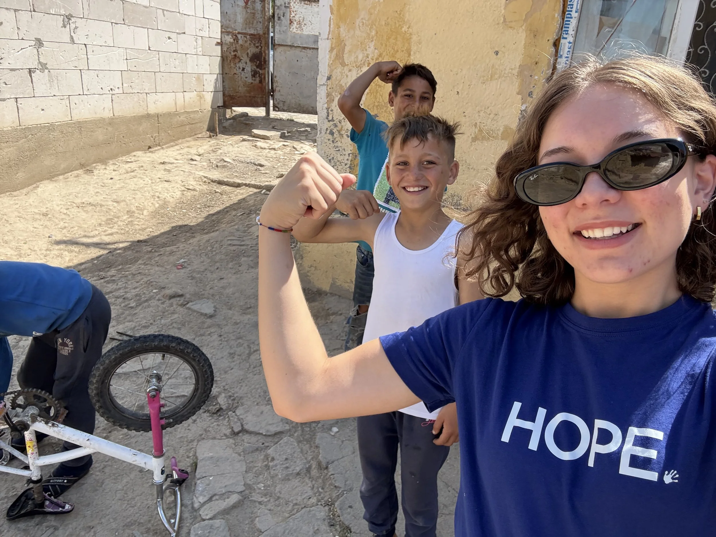 A woman wearing sunglasses and a blue shirt with the word 'HOPE' written on it is taking a selfie with three young boys smiling and flexible outside in a sunny, outdoor environment.