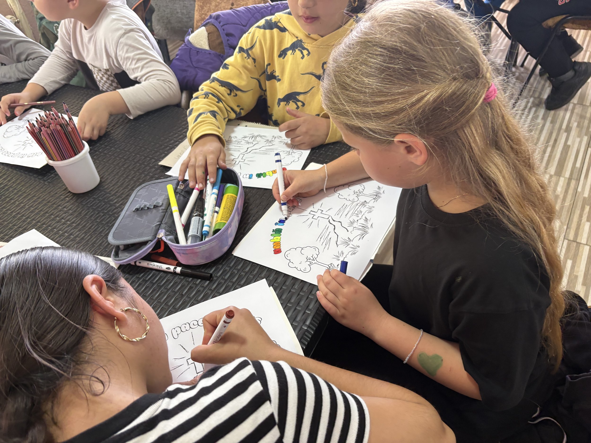 Children sitting at a table coloring pictures with markers, with coloring supplies in containers nearby.