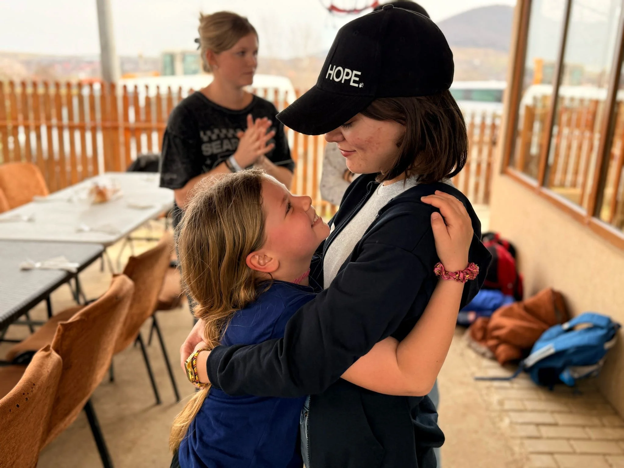 A young girl and a woman are hugging and smiling at each other inside a room with large windows. The girl has long blonde hair and is wearing a blue jacket, while the woman has dark hair, a black hat with the word 'HOPE' on it, and a black jacket. In