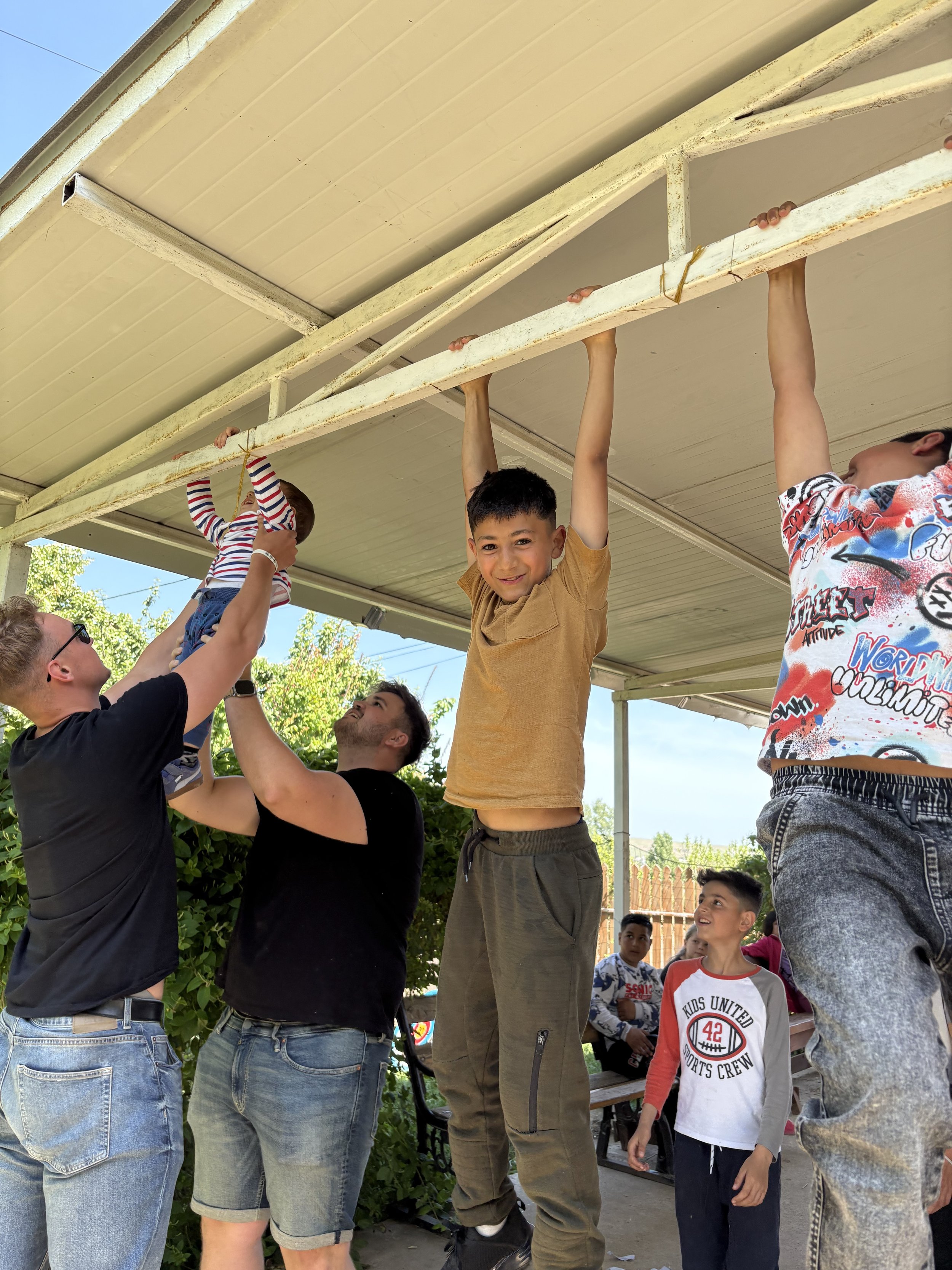 Kids playing on monkey bars under a covered outdoor area, with some children hanging and others watching and waiting.