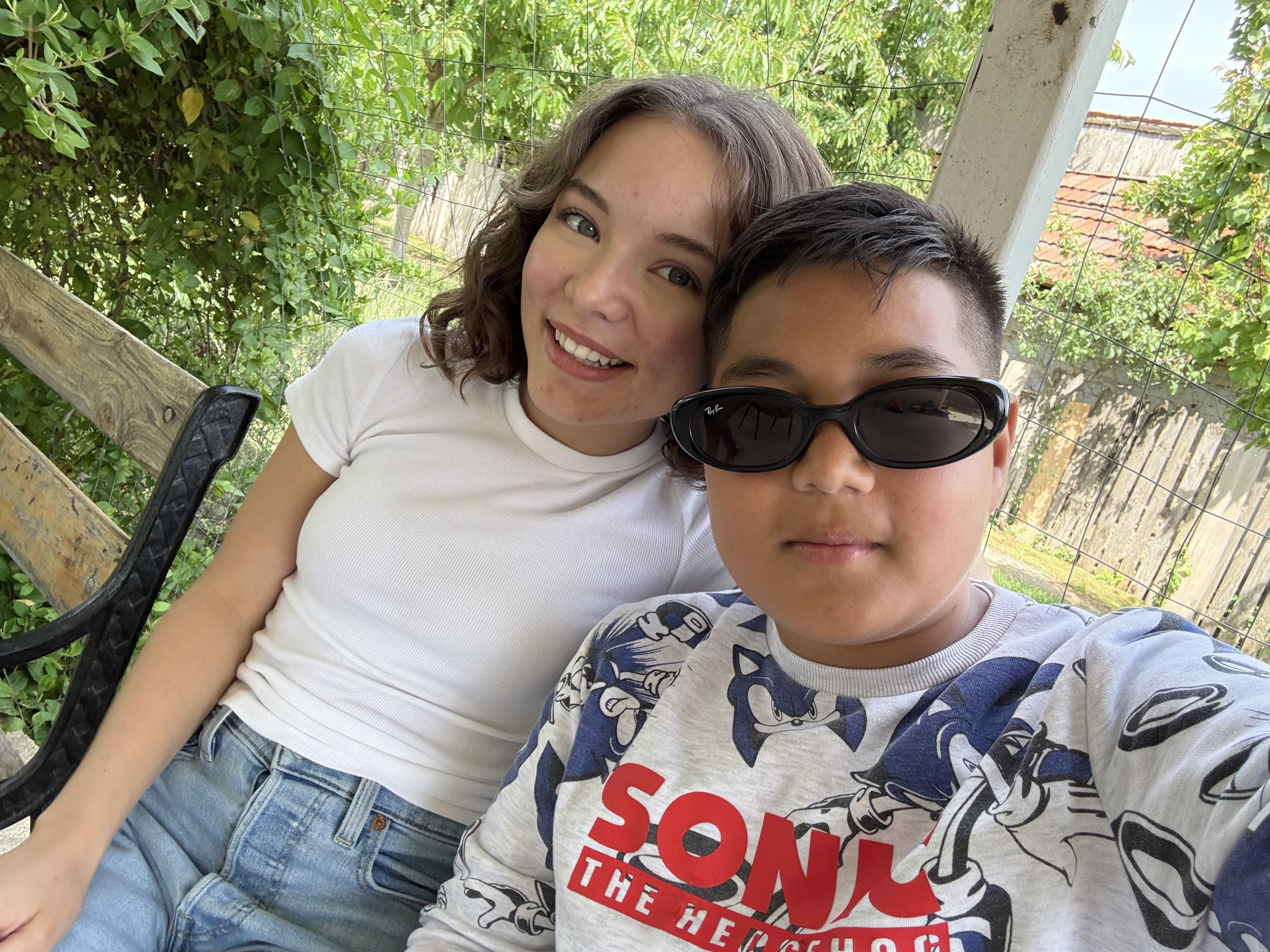 A smiling young woman and a boy taking a selfie outdoors on a bench surrounded by greenery. The woman has curly hair and is wearing a white t-shirt. The boy is wearing sunglasses and a Sonic the Hedgehog t-shirt.