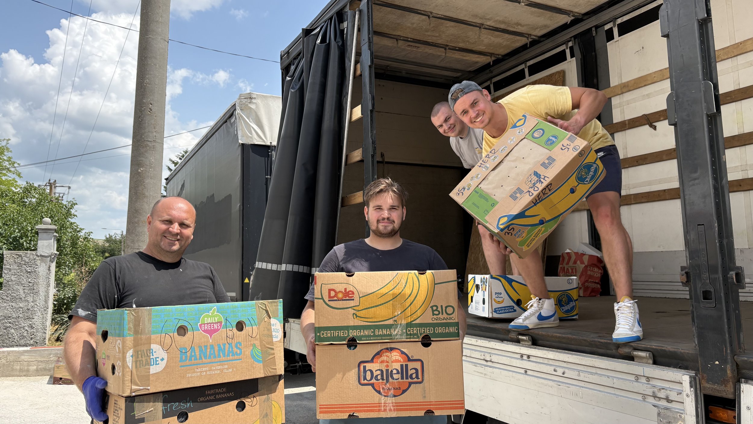 Four men unloading boxes of bananas from a truck, smiling at the camera. Two of them are standing outside the truck, holding boxes, while two are inside the truck, one holding a box.