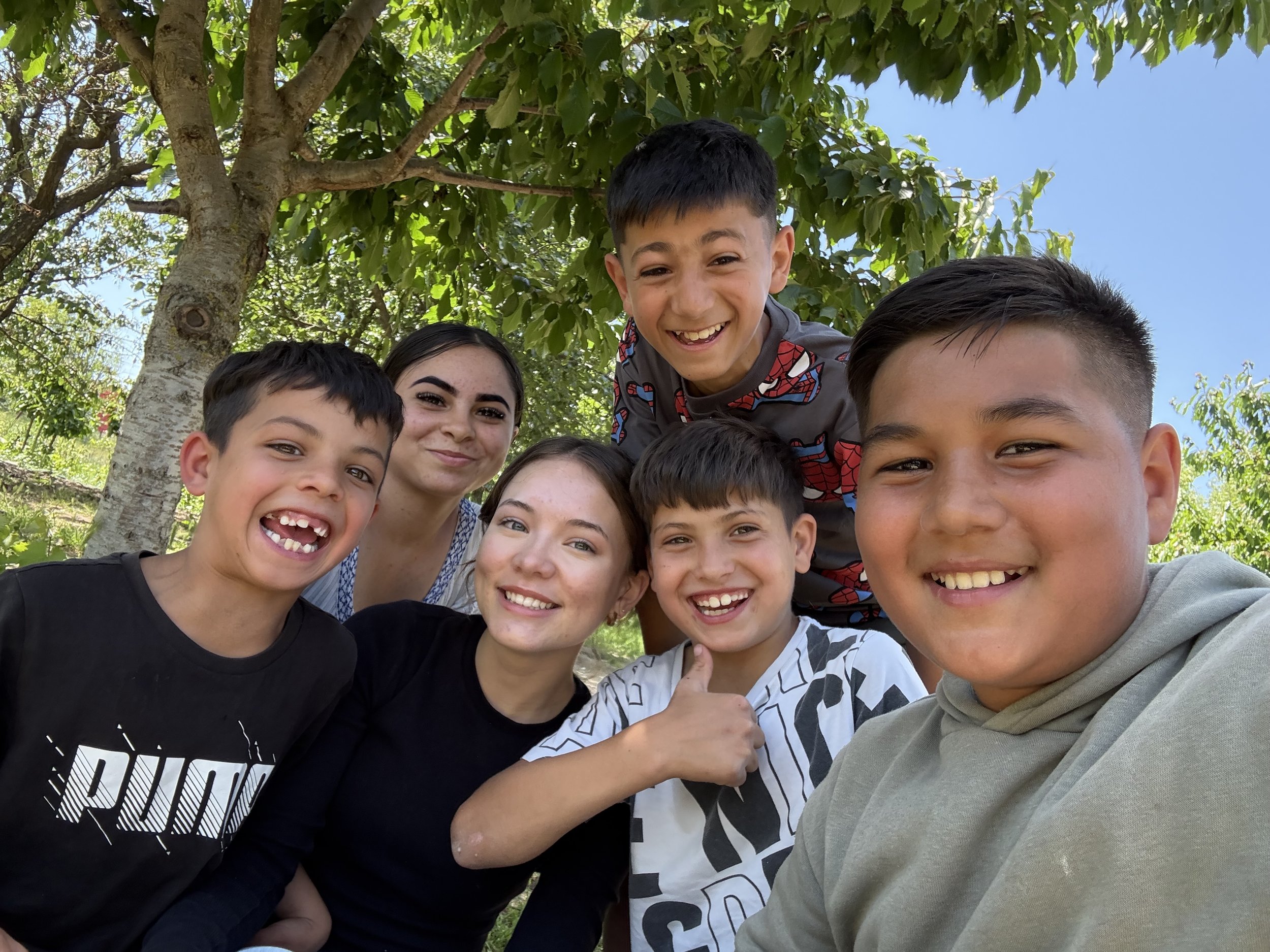 Group of seven children smiling and taking a selfie outdoors under a tree.