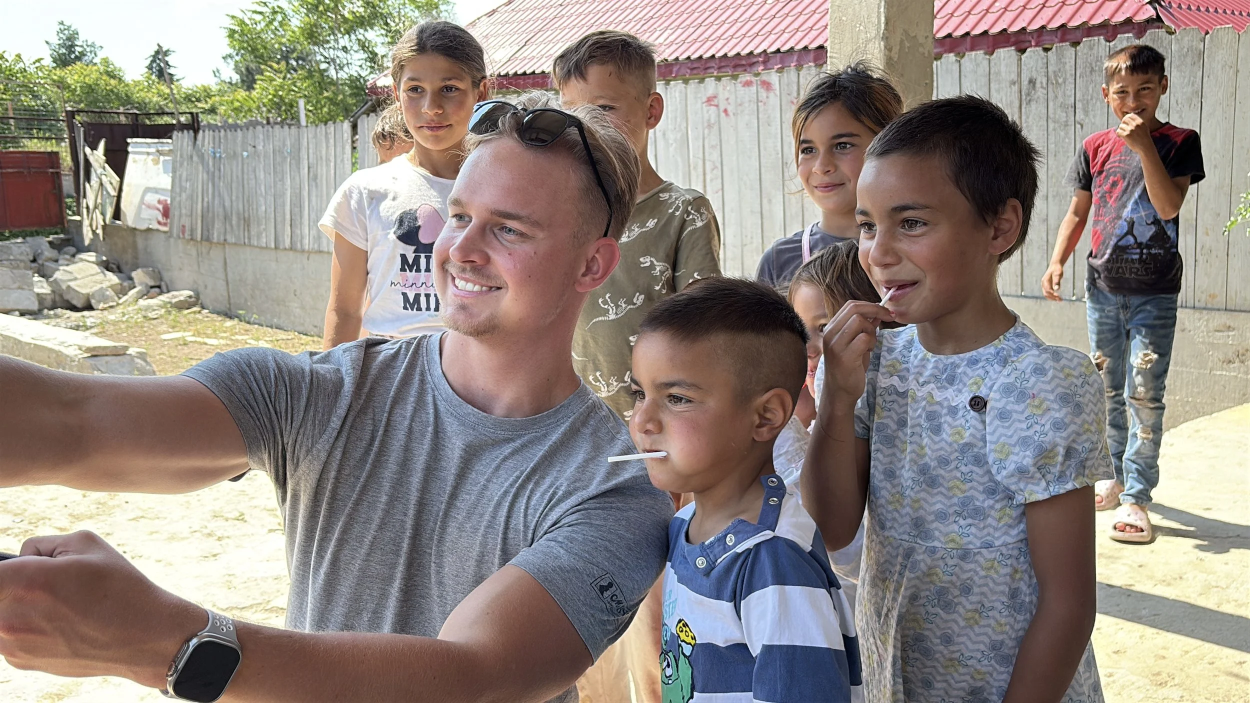 A man taking a selfie with a group of children outdoors, some children licking lollipops, in a sunny yard with a wooden fence and trees in the background.