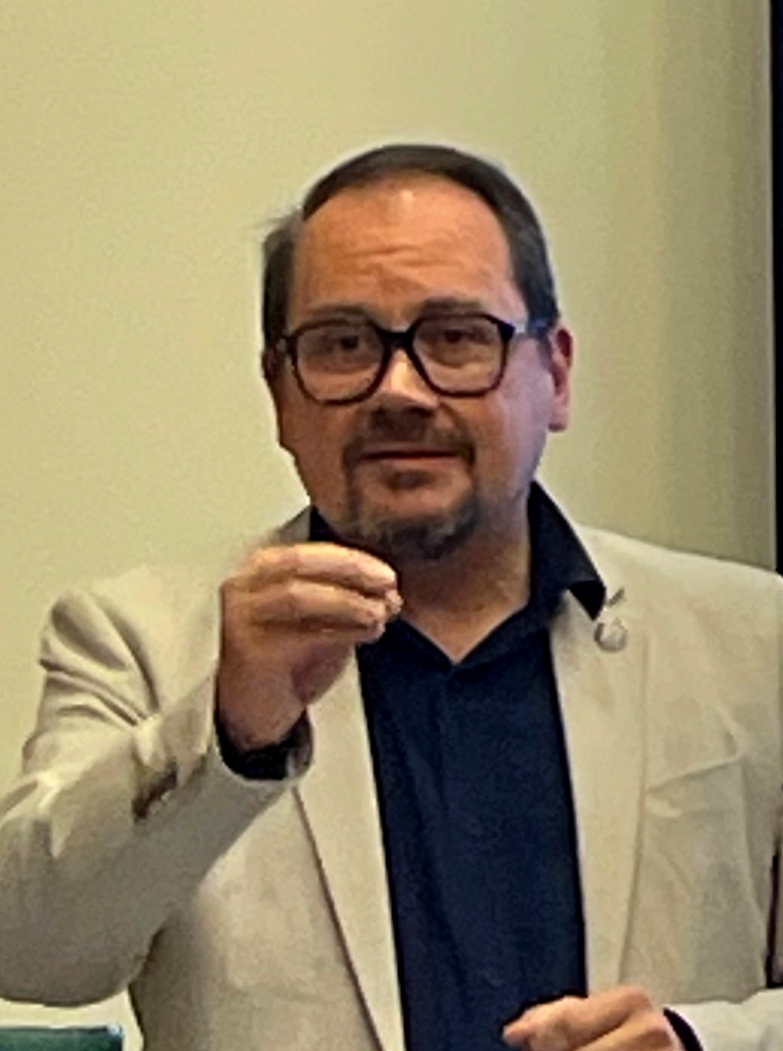 Un homme portant des lunettes, une veste beige et une chemise noire, parlant ou expliquant quelque chose.