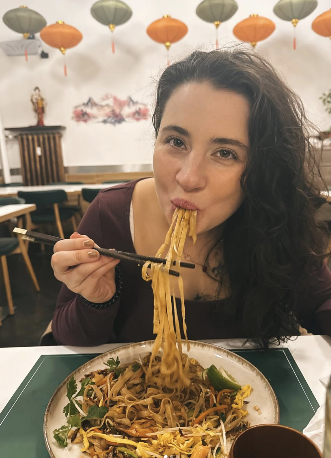 A woman with curly dark hair eating chopsticks of noodles in a restaurant with Asian decor and lanterns.