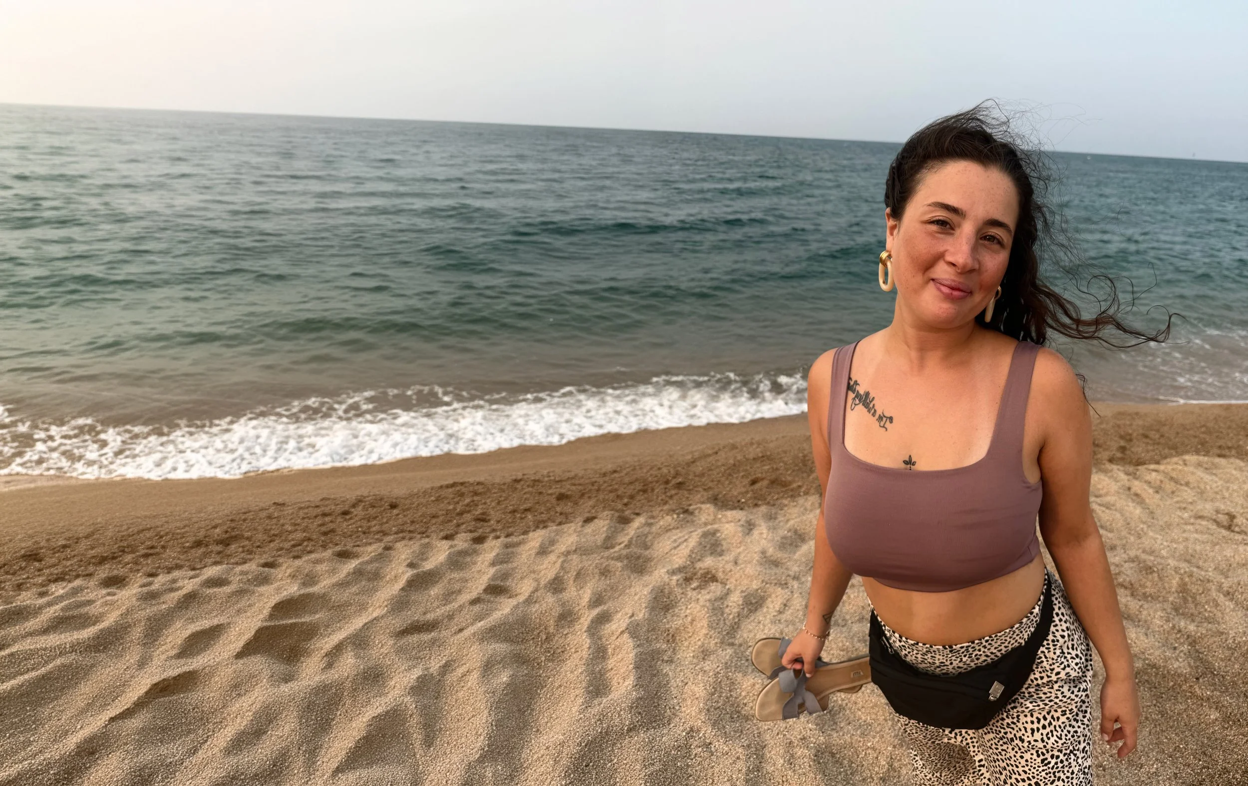 A woman with dark wavy hair, wearing large gold hoop earrings, a mauve crop top, and animal print pants, standing on a sandy beach with the ocean and a cloudy sky in the background, holding a pair of beige high heels in her right hand.