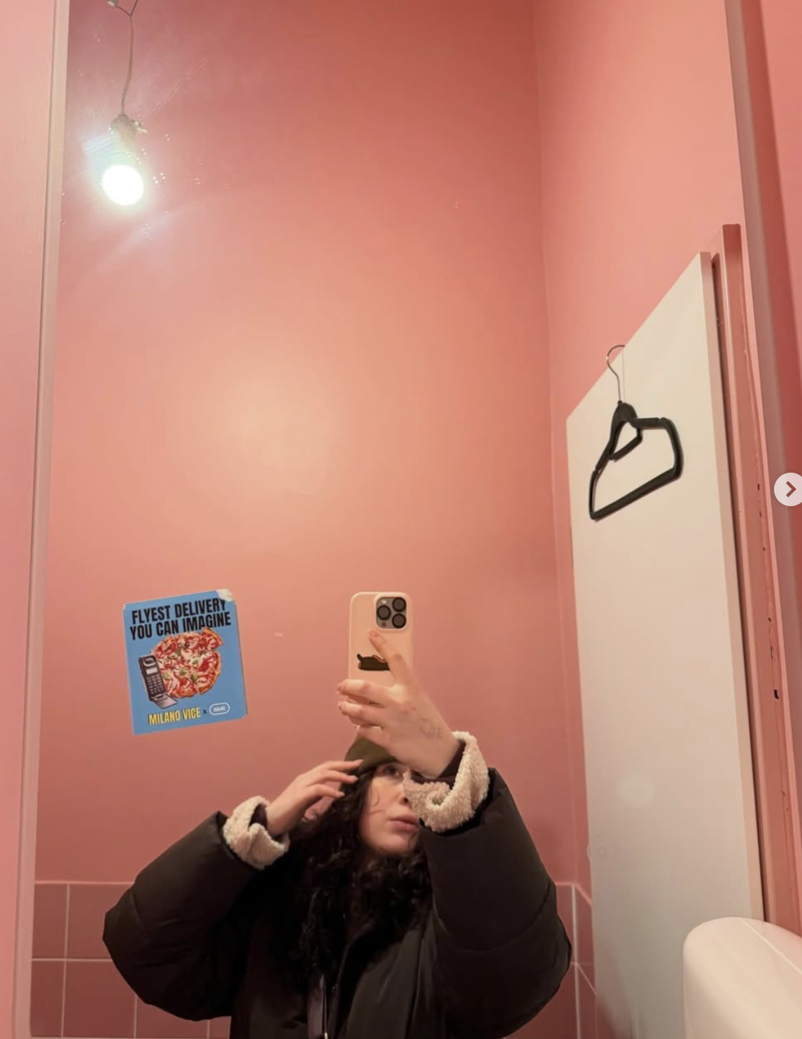 A woman taking a mirror selfie in a pink-colored bathroom with a lantern-style light, a black hanger on the wall, and a blue poster that reads "Flyest Delivery You Can Imagine" with a pizza illustration.